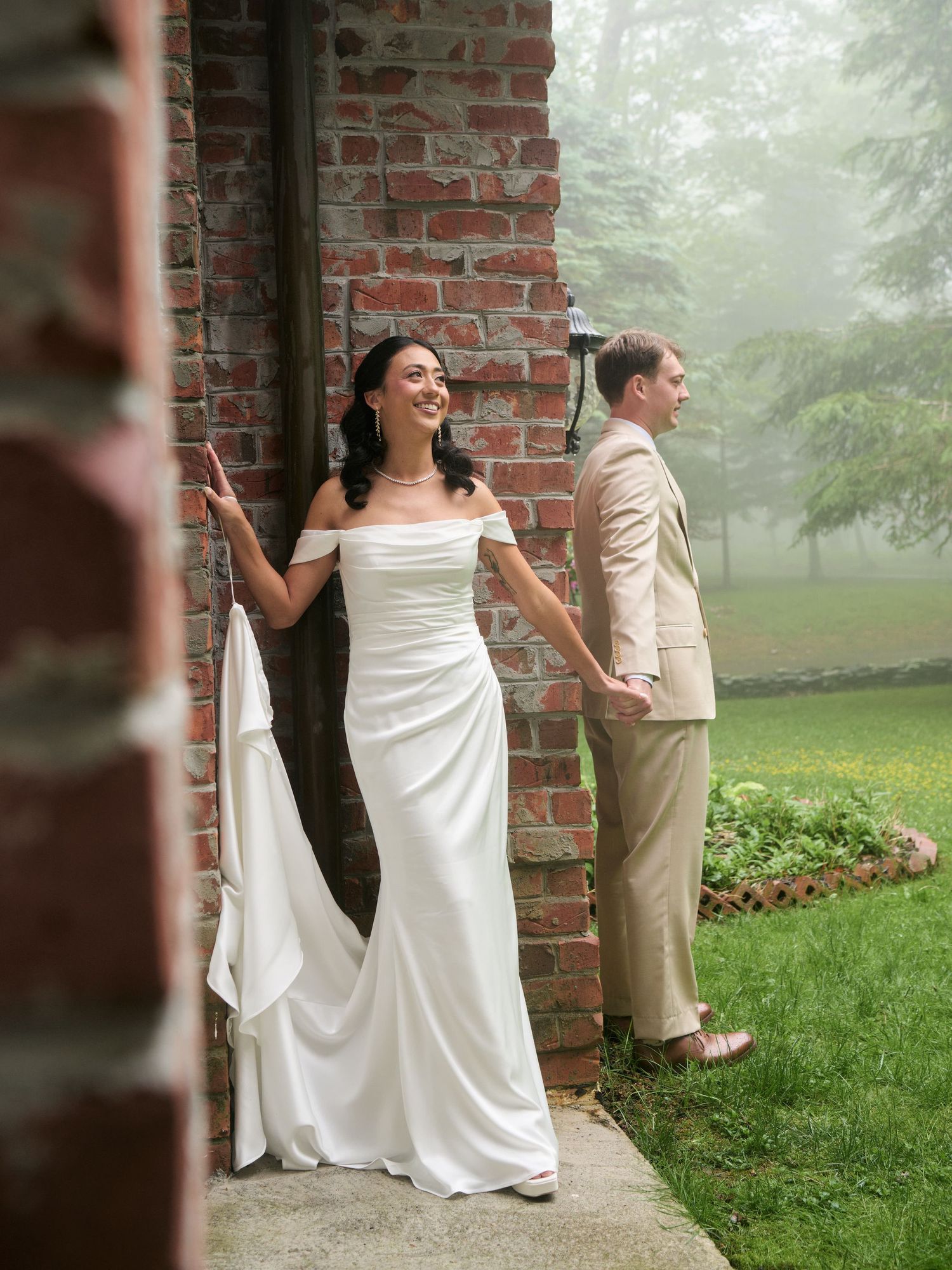 Bride in white off-shoulder gown and groom in tan suit pose by brick wall in misty outdoor wedding setting.