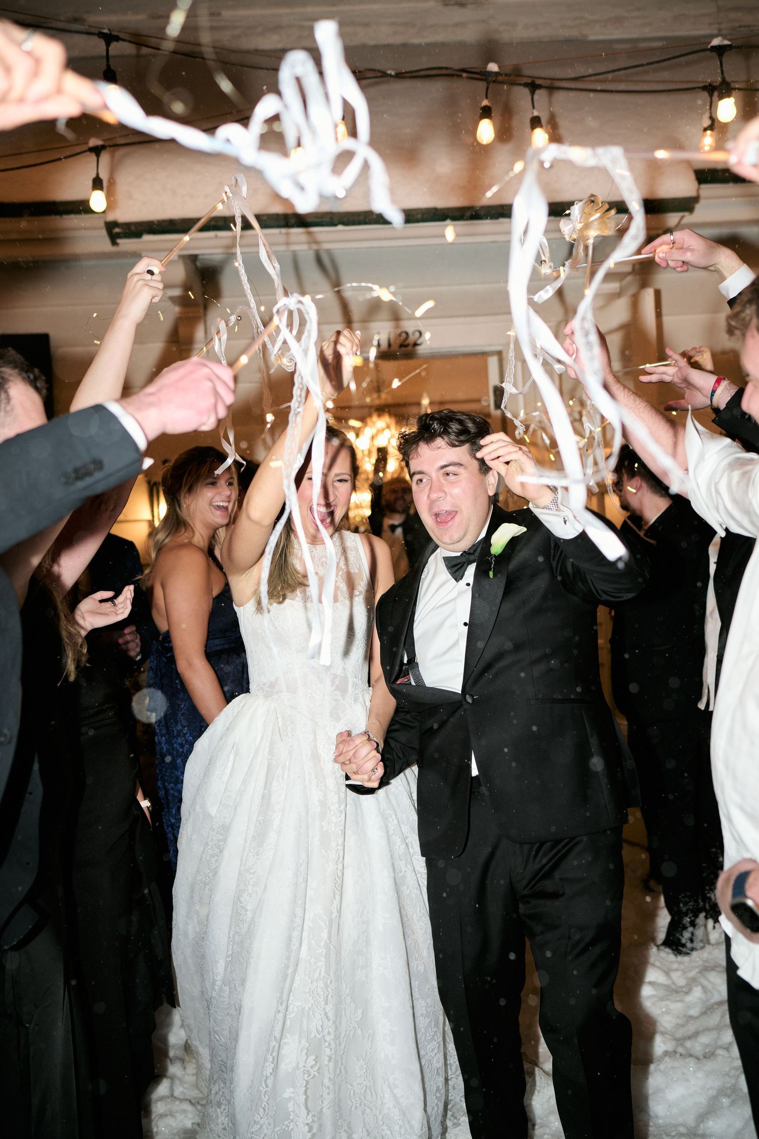 Bride and groom exit wedding reception through streamers while guests celebrate around them.