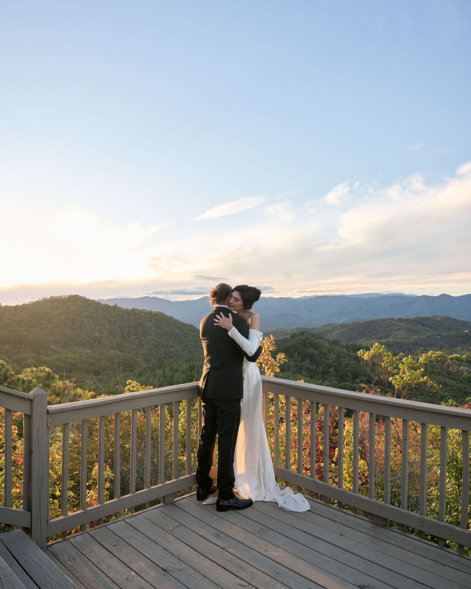 A couple shares a romantic moment on a mountain deck overlooking a scenic mountain range at sunset.