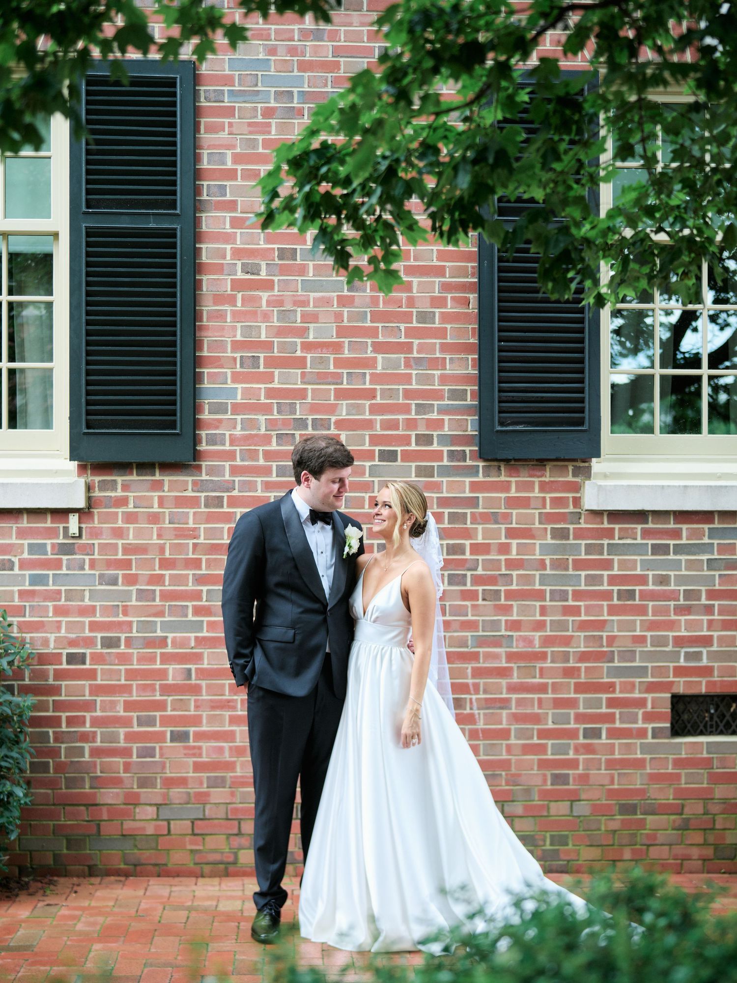A couple in wedding attire share a romantic moment against a red brick wall with black shutters and green foliage.