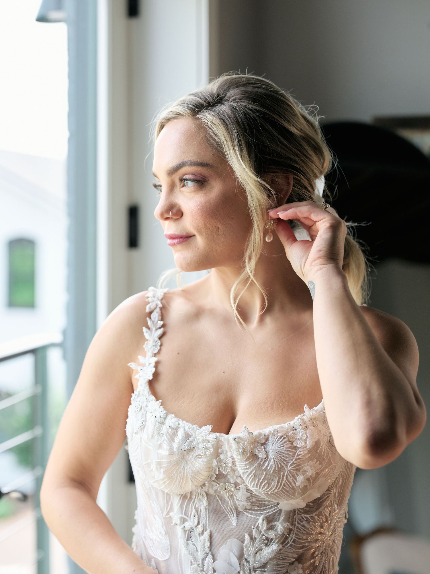 Bride wearing delicate white lace and floral dress adjusts earring while looking out window during wedding preparations.