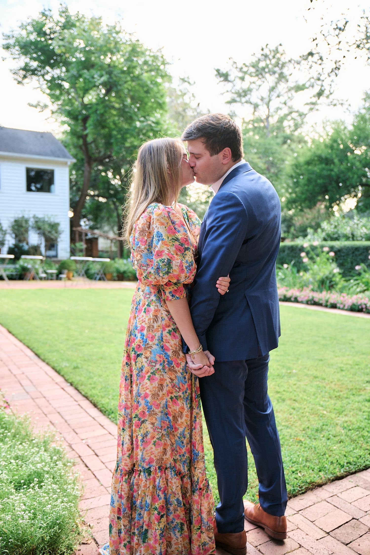Romantic couple embraces on brick path surrounded by lush garden landscape.