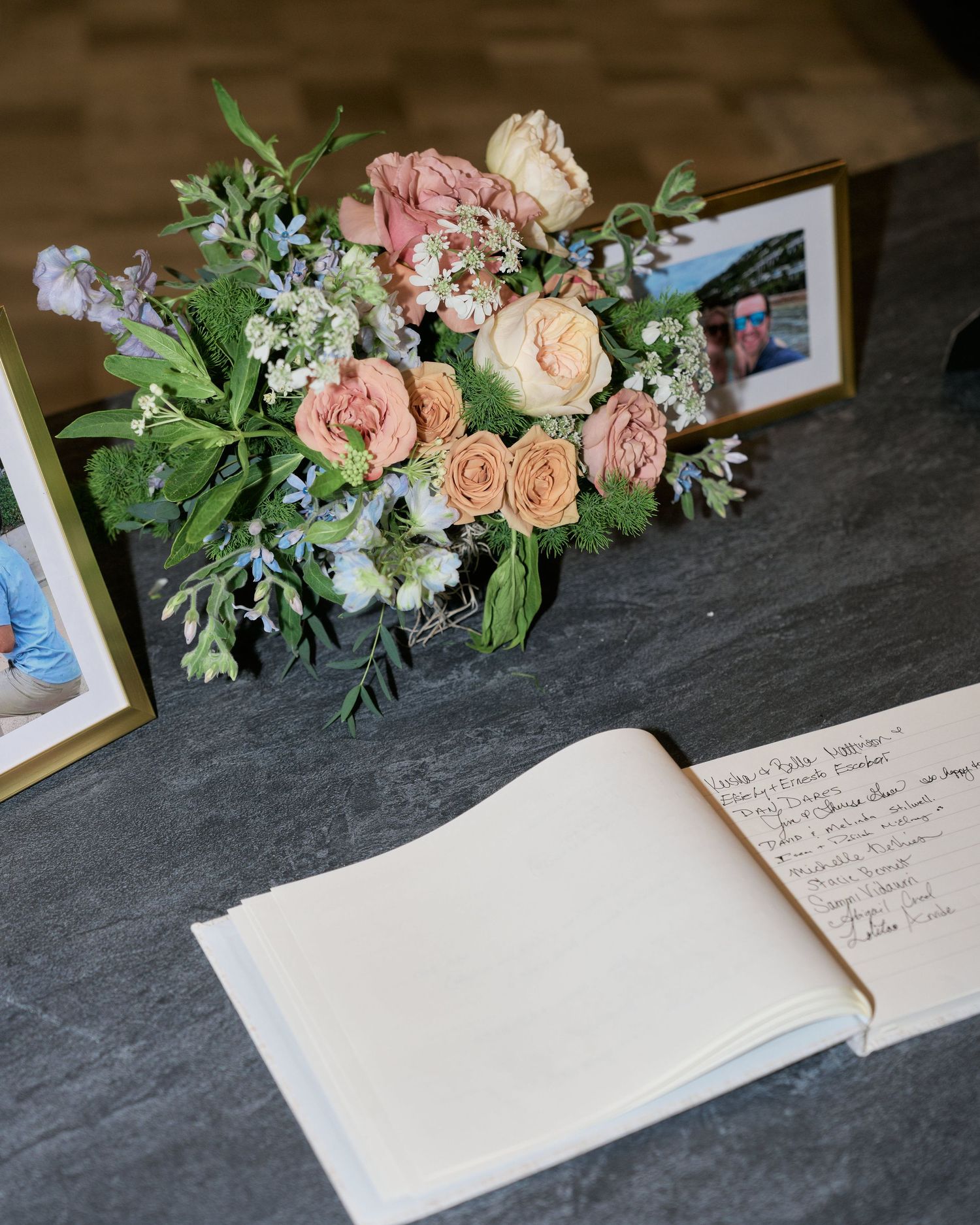 An elegant floral arrangement with pink roses and greenery next to an open guestbook on a dark surface.