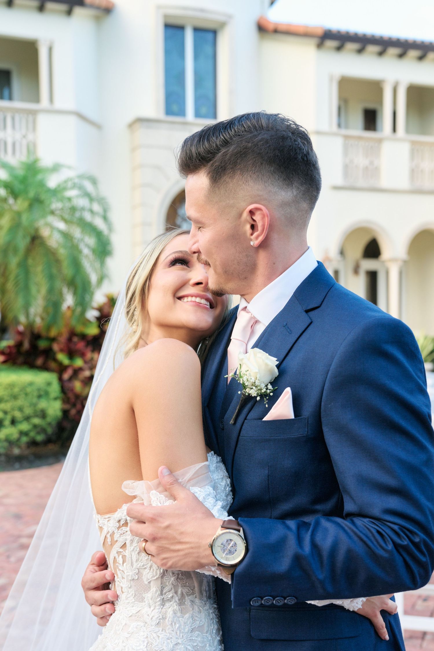 Intimate wedding portrait with couple embracing against Mediterranean villa backdrop.