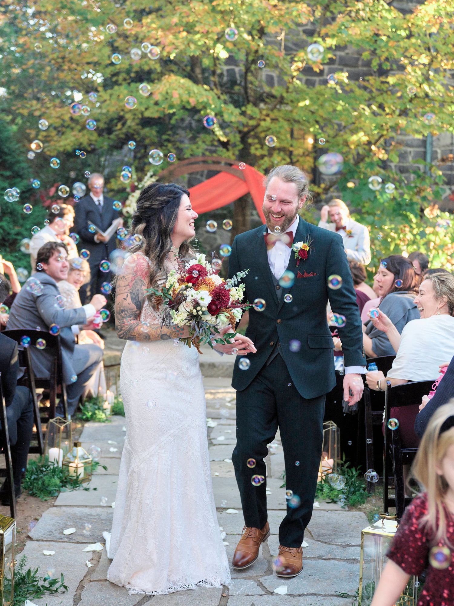 Newlyweds walking down aisle while guests blow bubbles during outdoor ceremony.