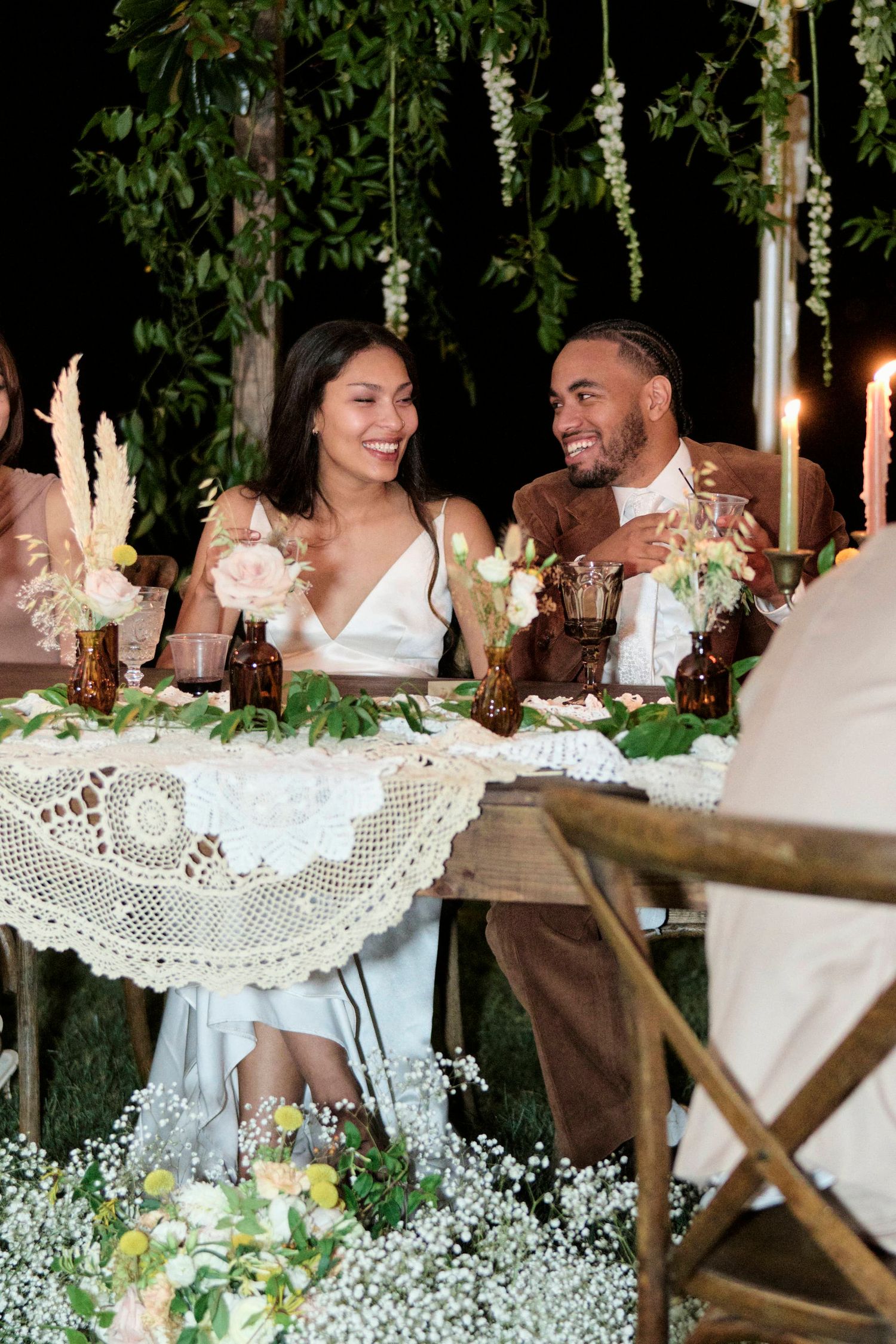 Newlyweds sharing a joyful moment at their reception table decorated with flowers and lace.