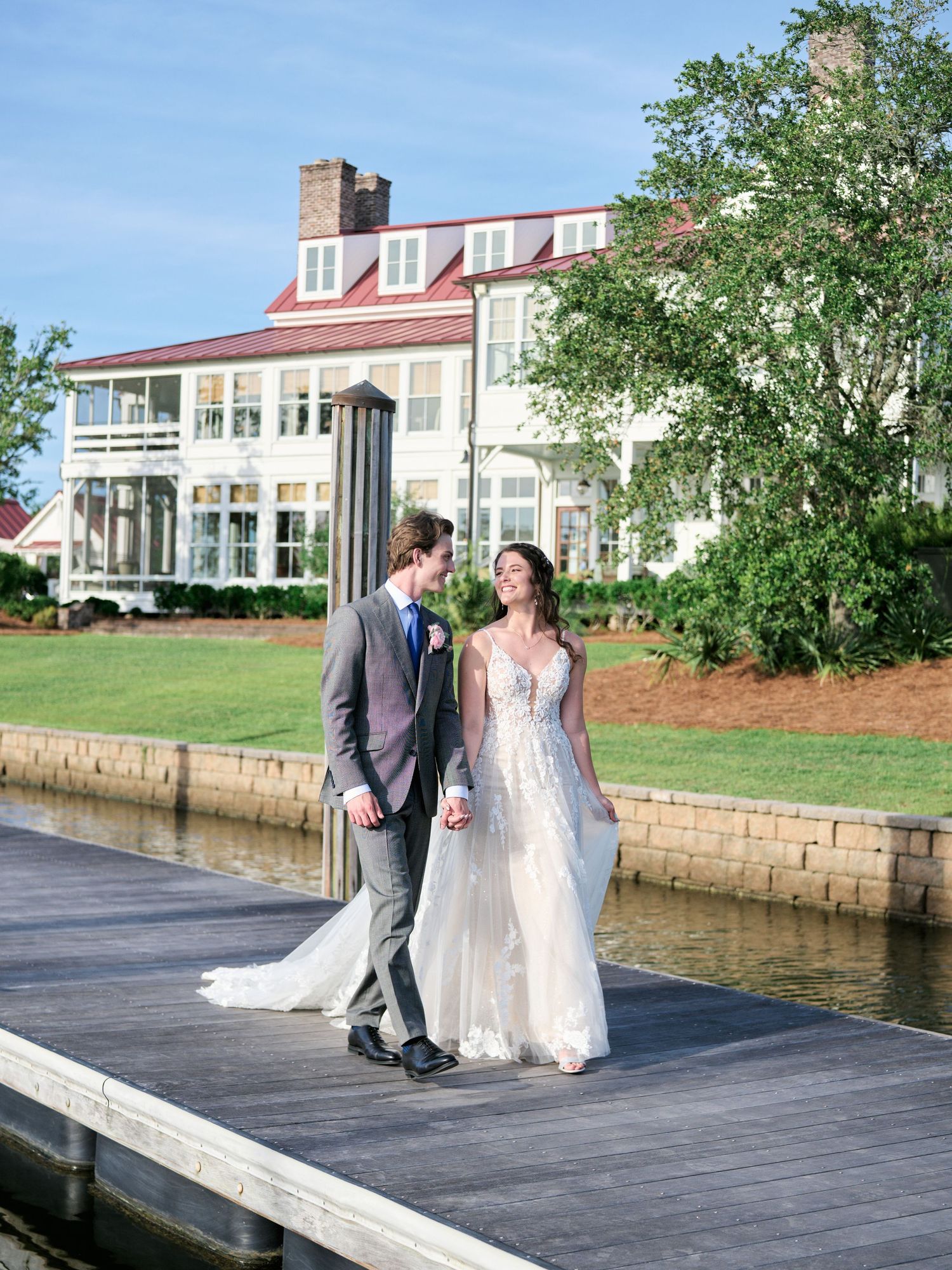 Couple walking along a waterfront dock with a grand mansion in the background.