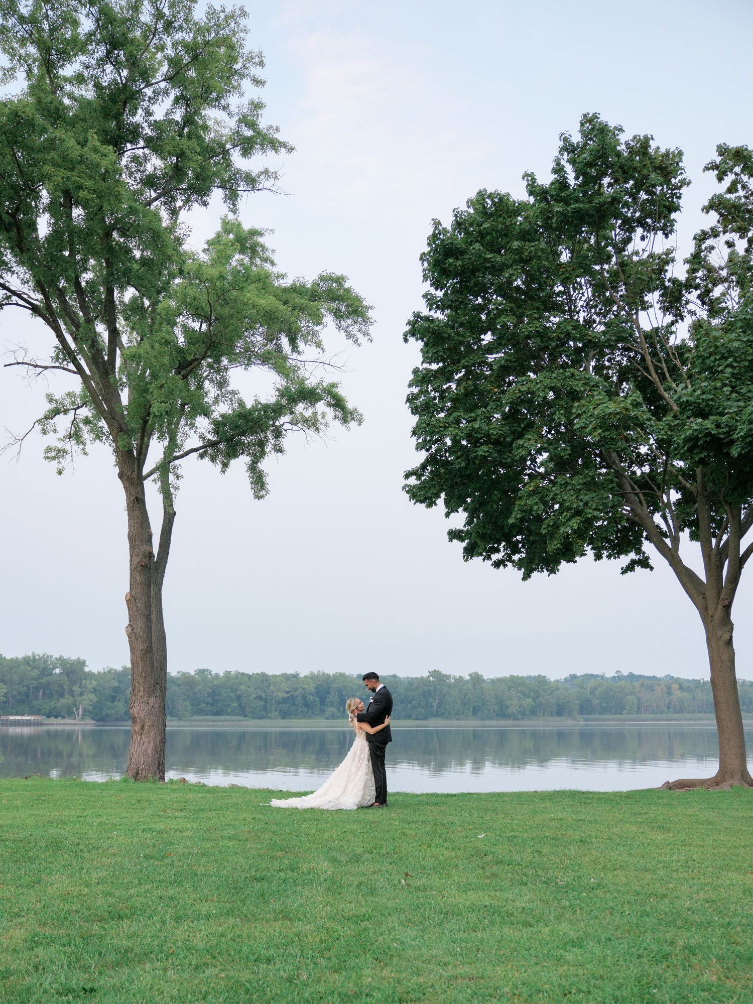 A couple shares a romantic moment between two tall trees on a grassy lawn overlooking a serene lake.
