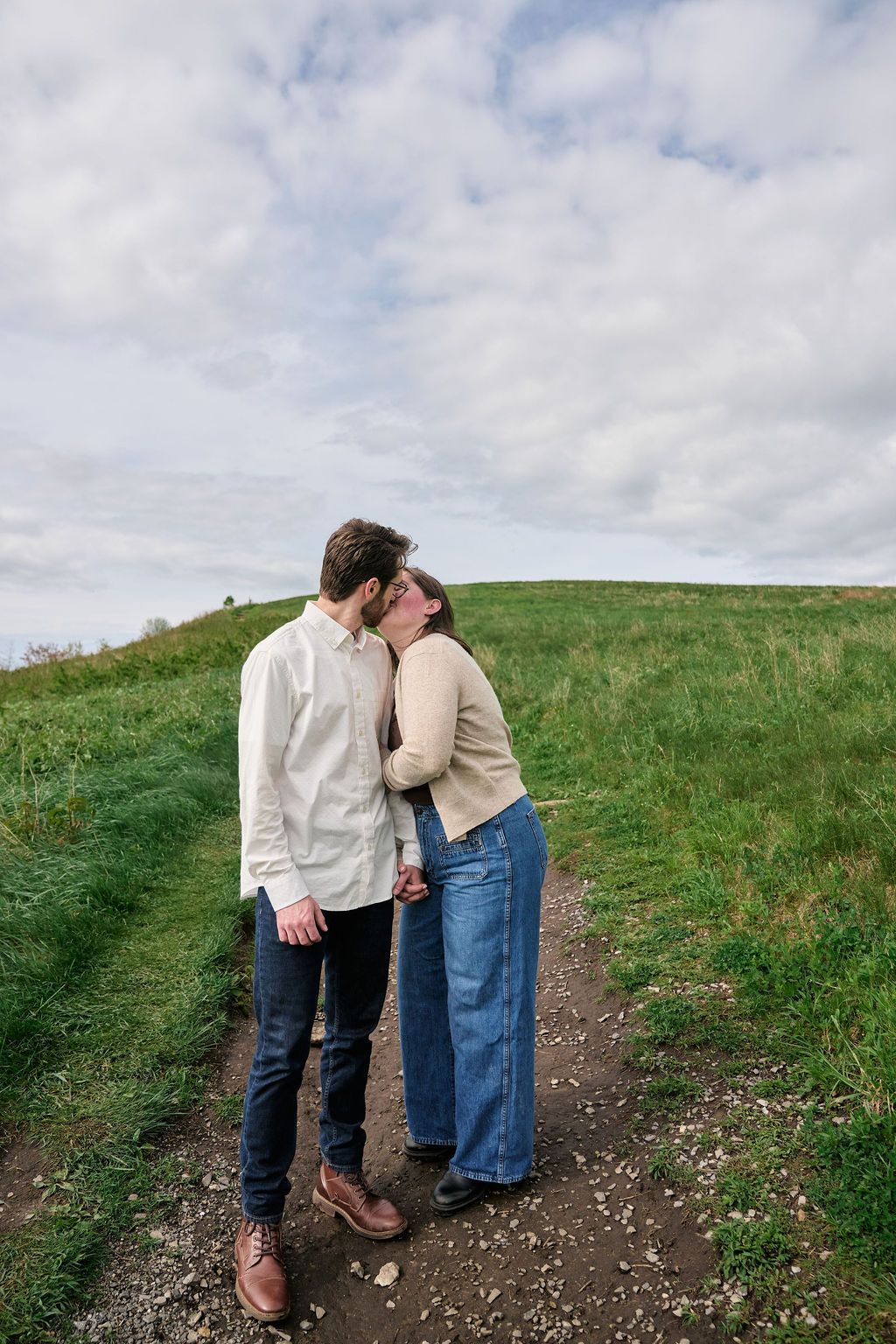 A couple shares a tender kiss while standing on a dirt path along a green grassy hillside under cloudy skies.