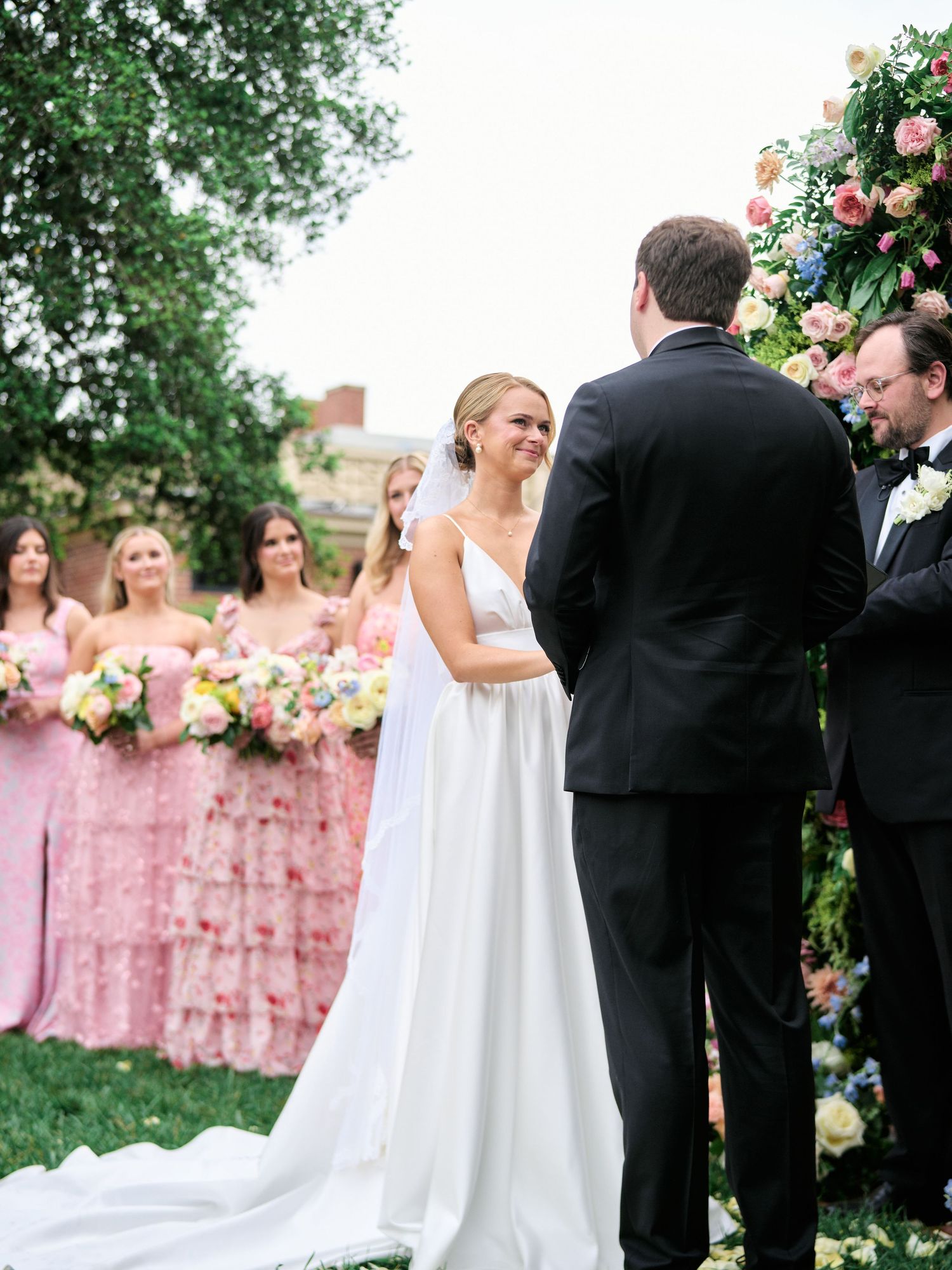 Wedding ceremony with bridesmaids in pink dresses standing beside a floral arch in a garden setting.