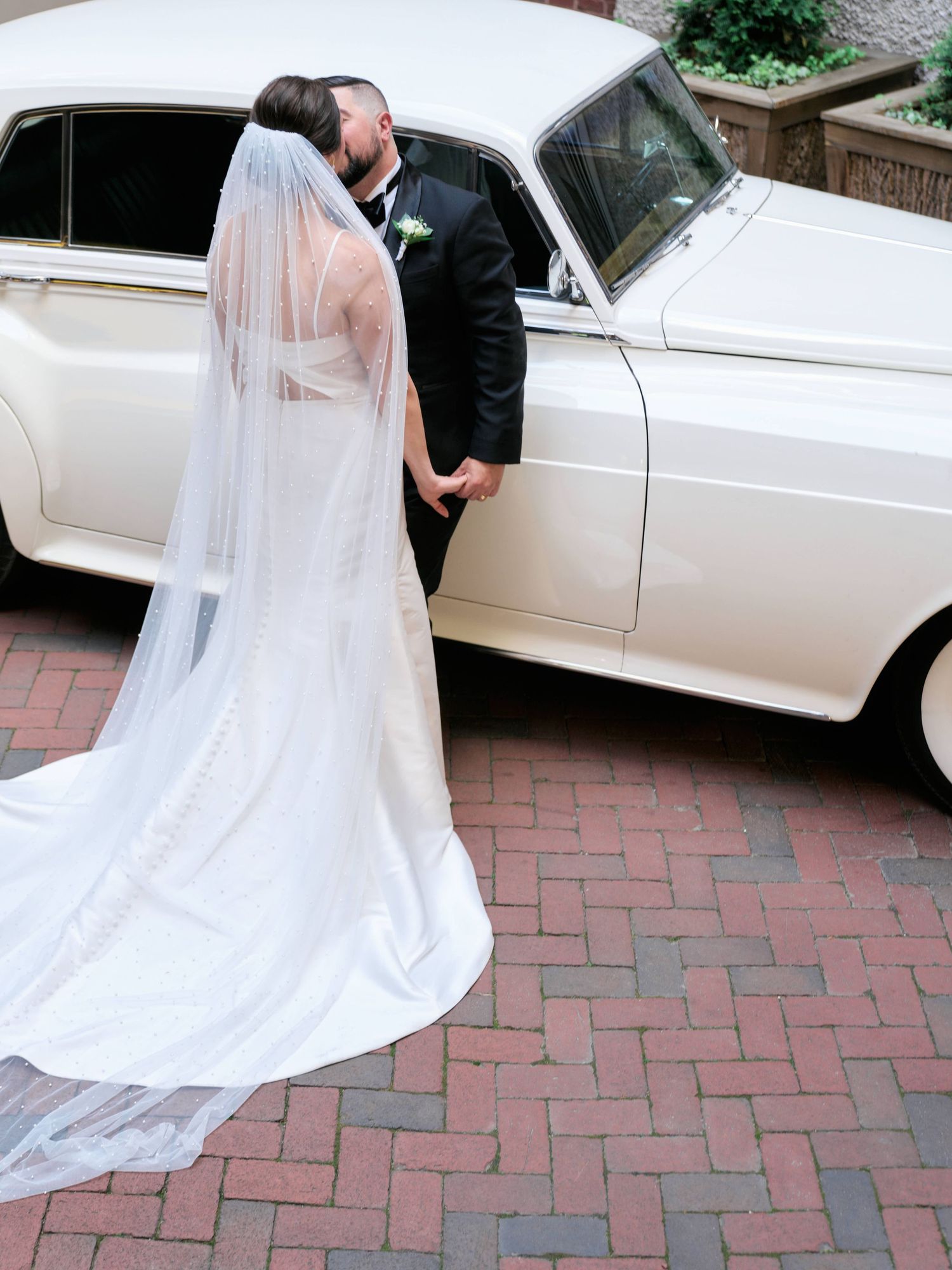 A flowing white wedding dress and veil twirl elegantly on a brick courtyard next to a classic vintage white car.