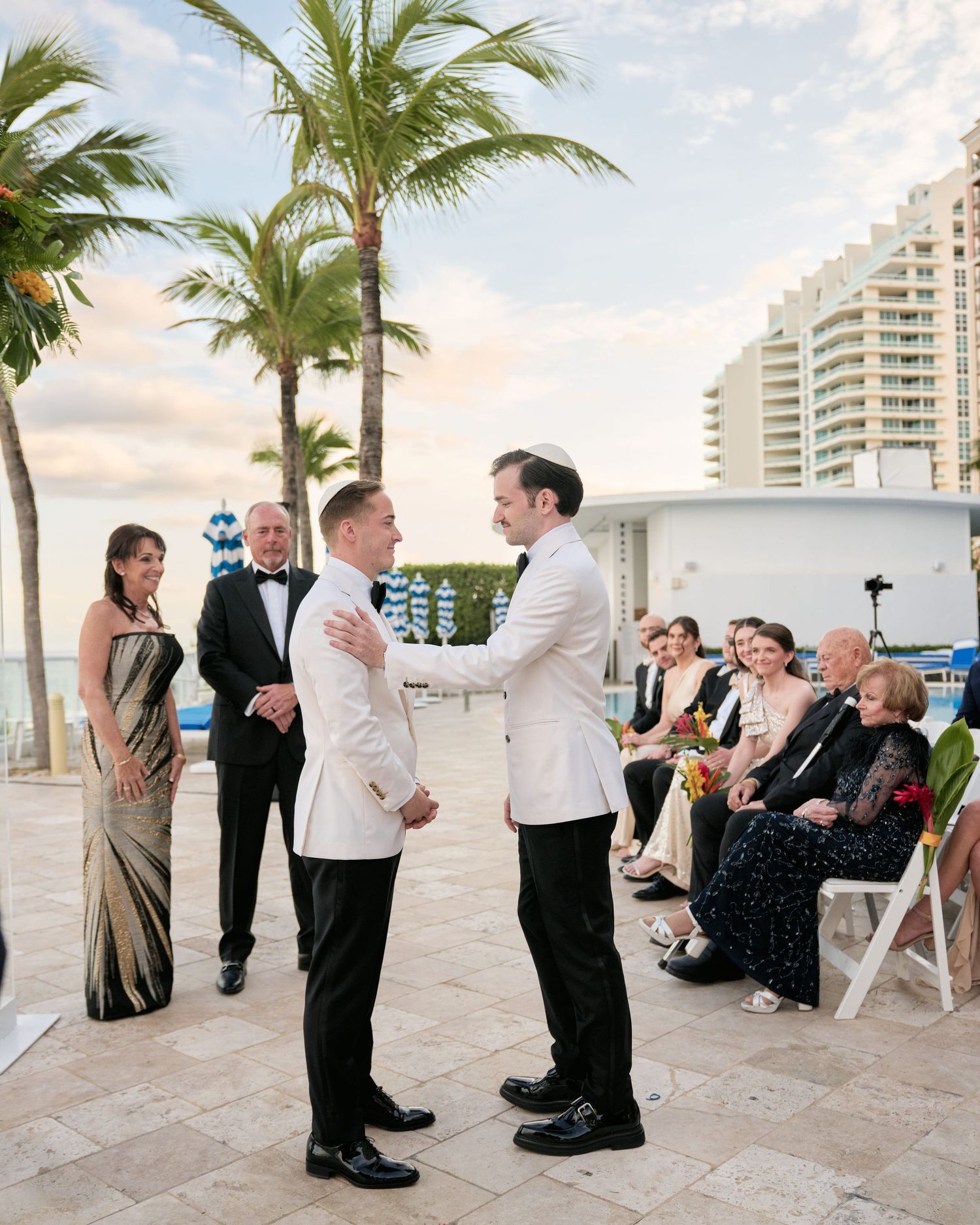 Same-sex wedding ceremony taking place at sunset on a beachfront with palm trees and high-rise buildings in background.