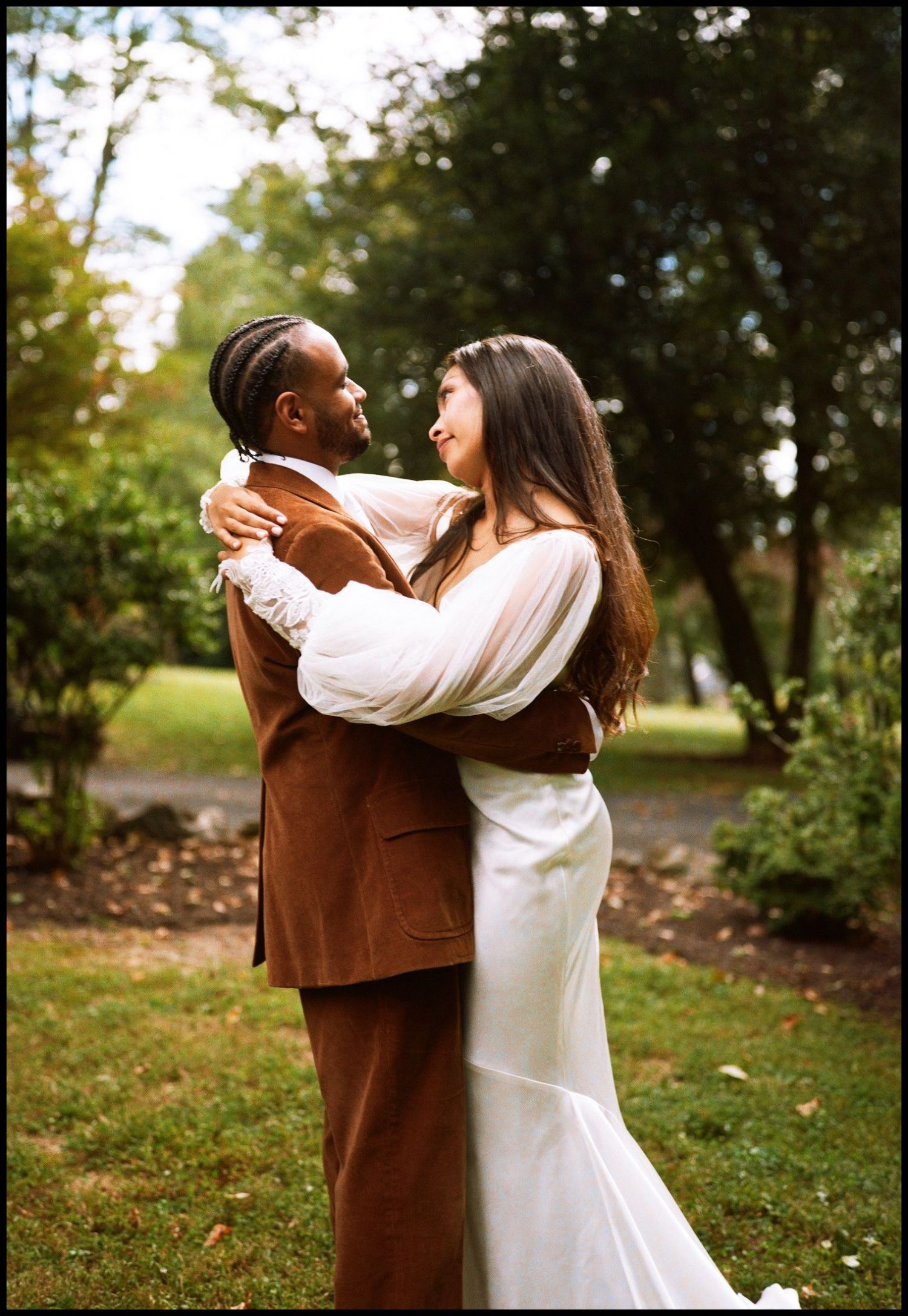 Happy couple embracing outdoors with bride in white dress and groom in brown suit.