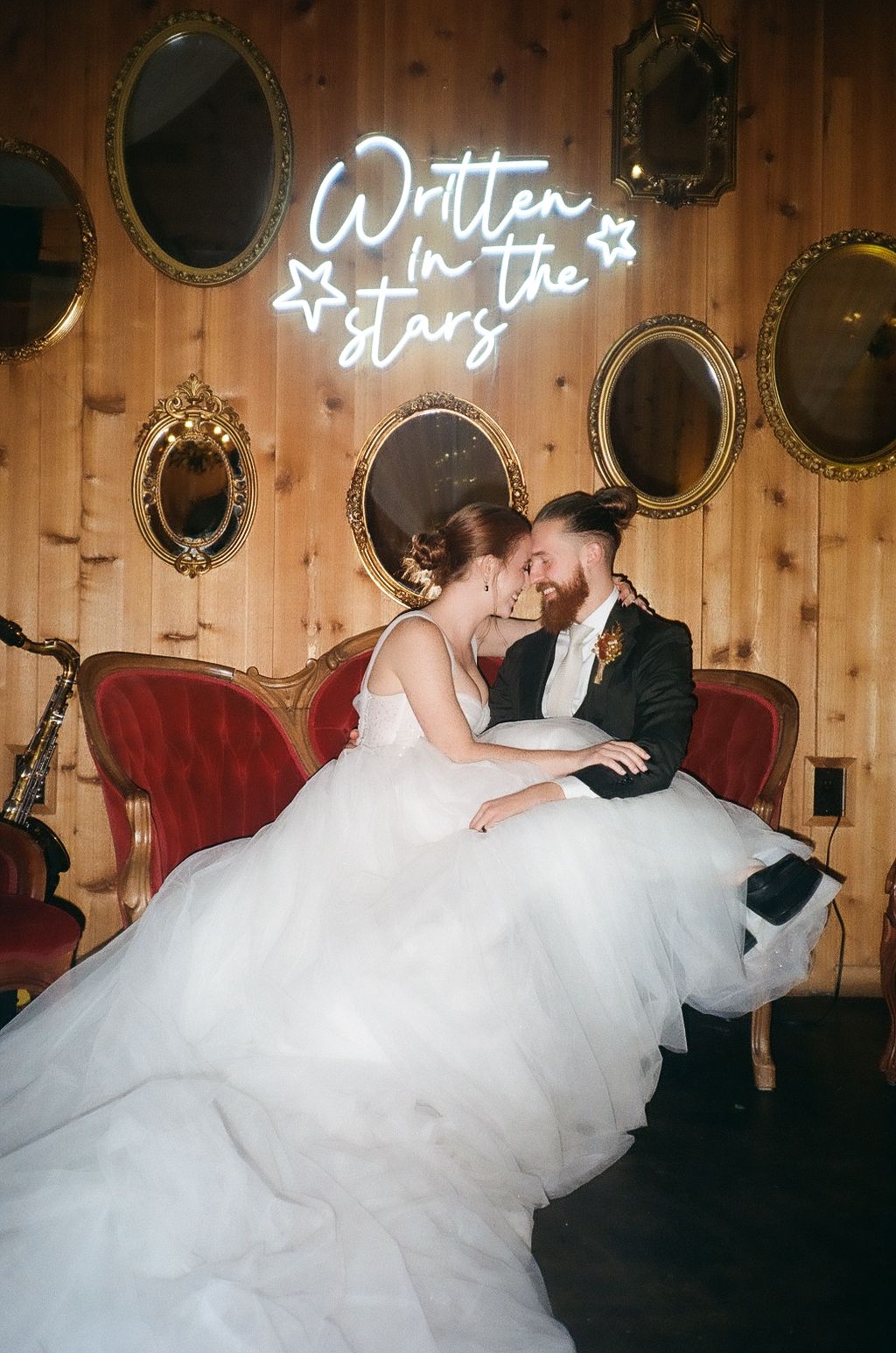 Newlyweds sharing intimate moment on red velvet couch under neon sign and vintage mirrors.