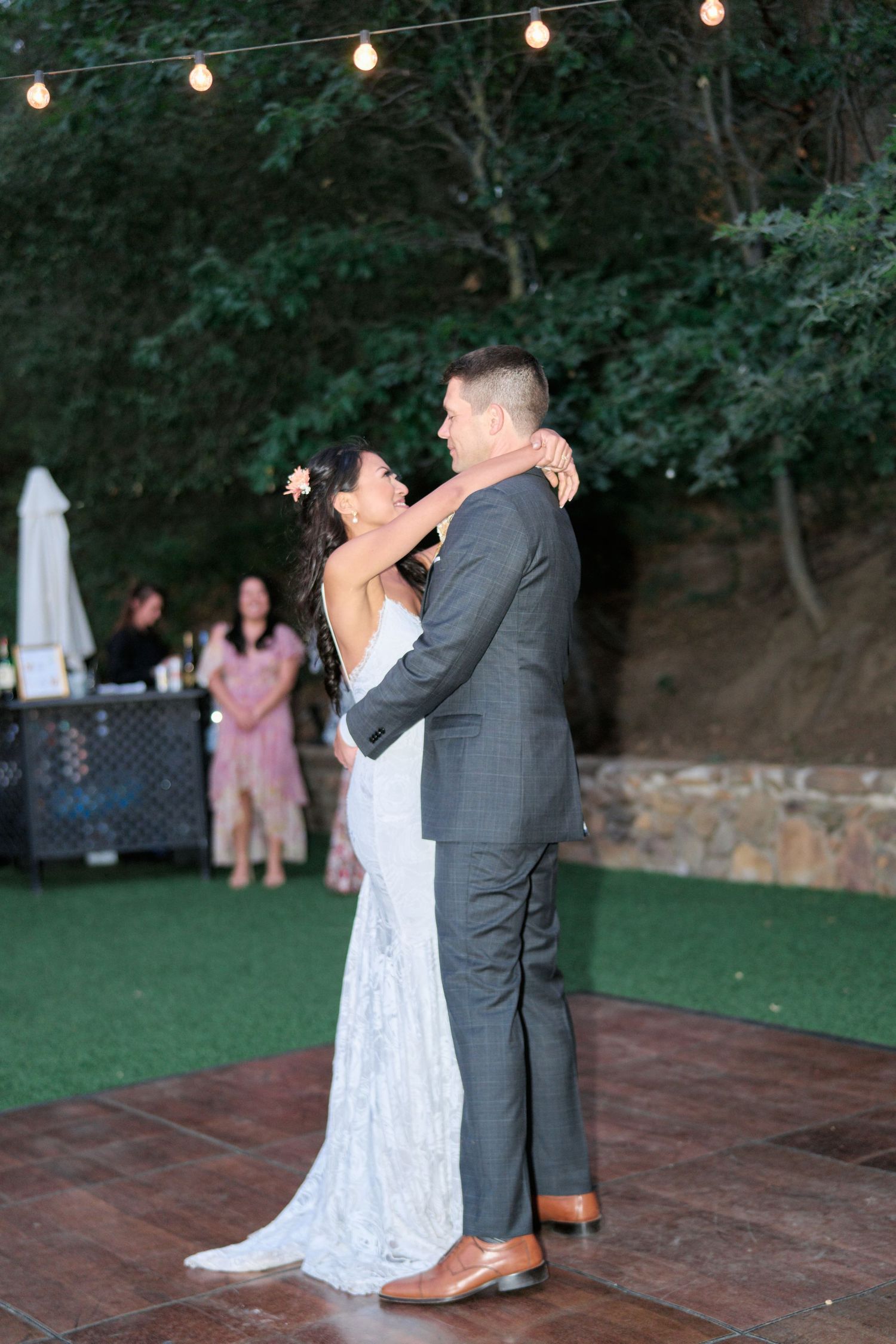 A couple dances on an outdoor dance floor decorated with string lights and greenery.