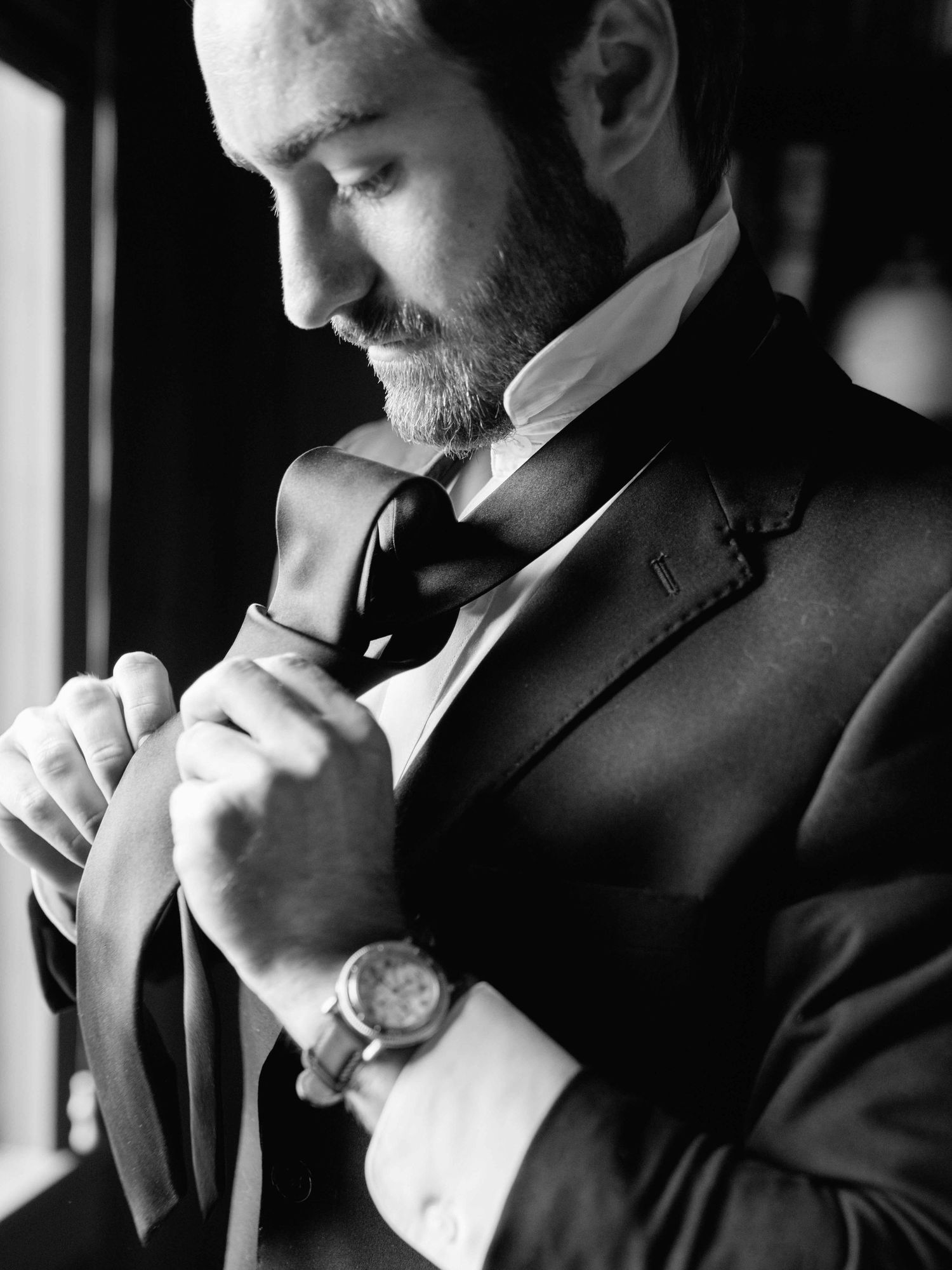 Black and white photo of a groom in a tuxedo adjusting his bow tie, wearing a watch, preparing for his wedding.