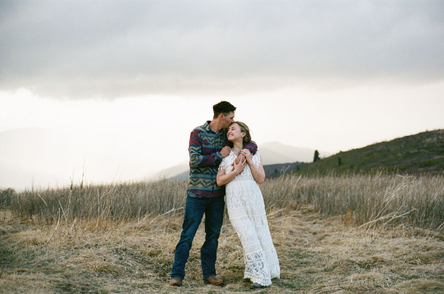 A couple in wedding attire shares an intimate moment in a scenic field of tall grass at sunset.