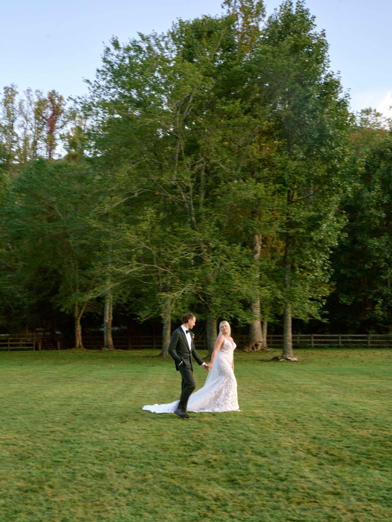 A couple in wedding attire stands on a lush green lawn with tall trees creating a natural backdrop at dusk.