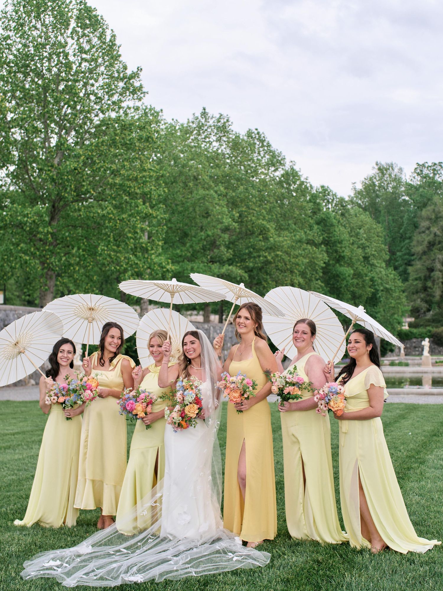 Wedding party in yellow dresses pose with white parasols on a grassy lawn during a summer celebration.
