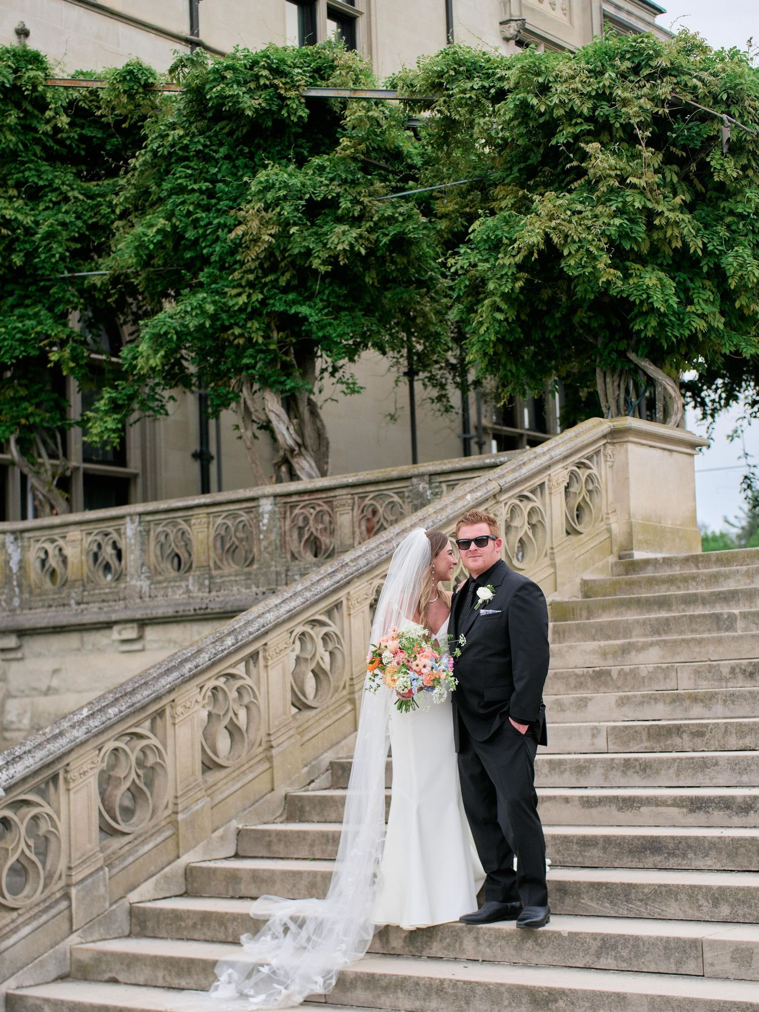 A romantic wedding couple poses on elegant stone steps with ornate railings and lush greenery overhead.