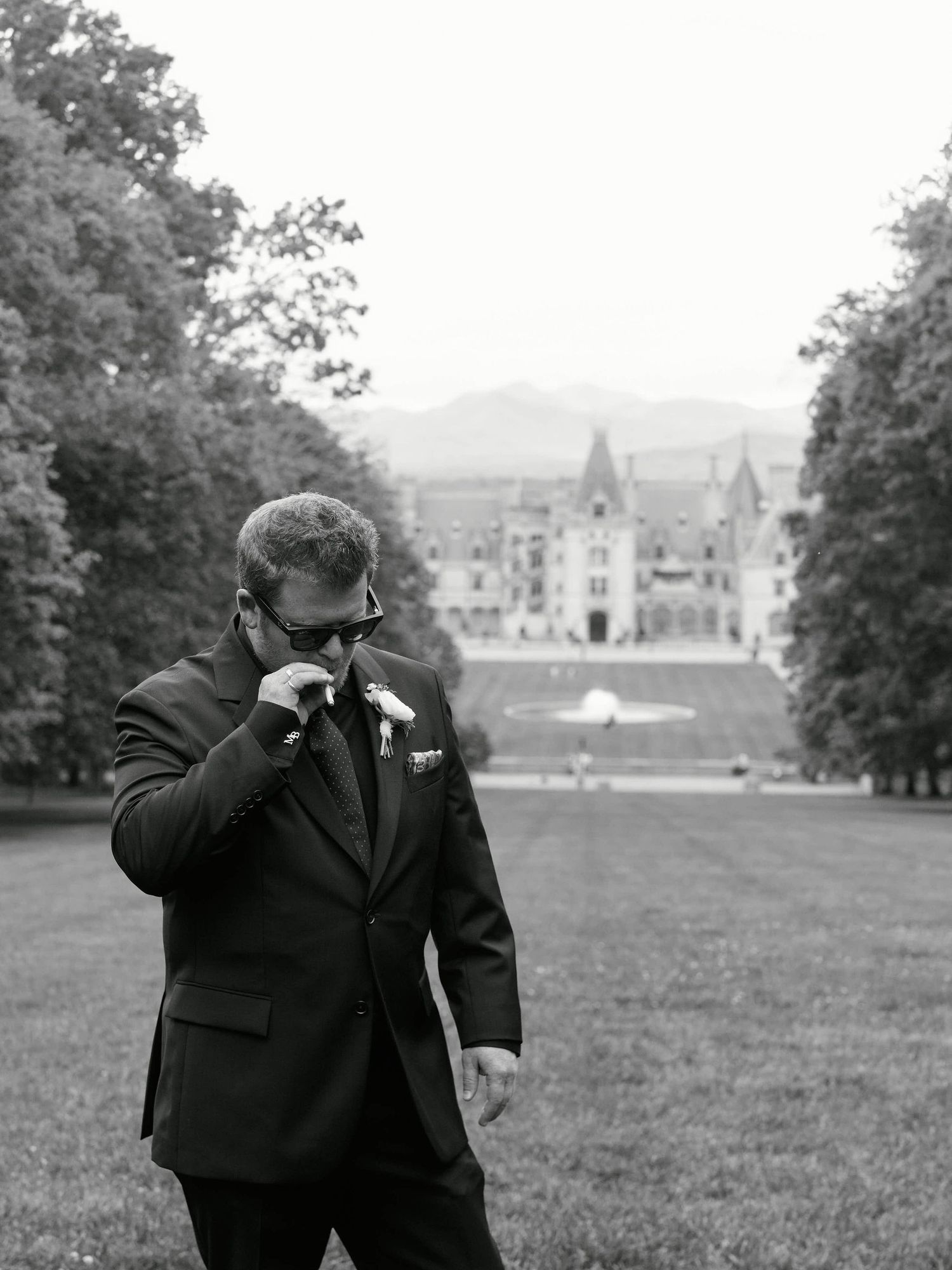 A black and white photo of two figures in formal attire walking together across a grassy field with buildings in the distance.