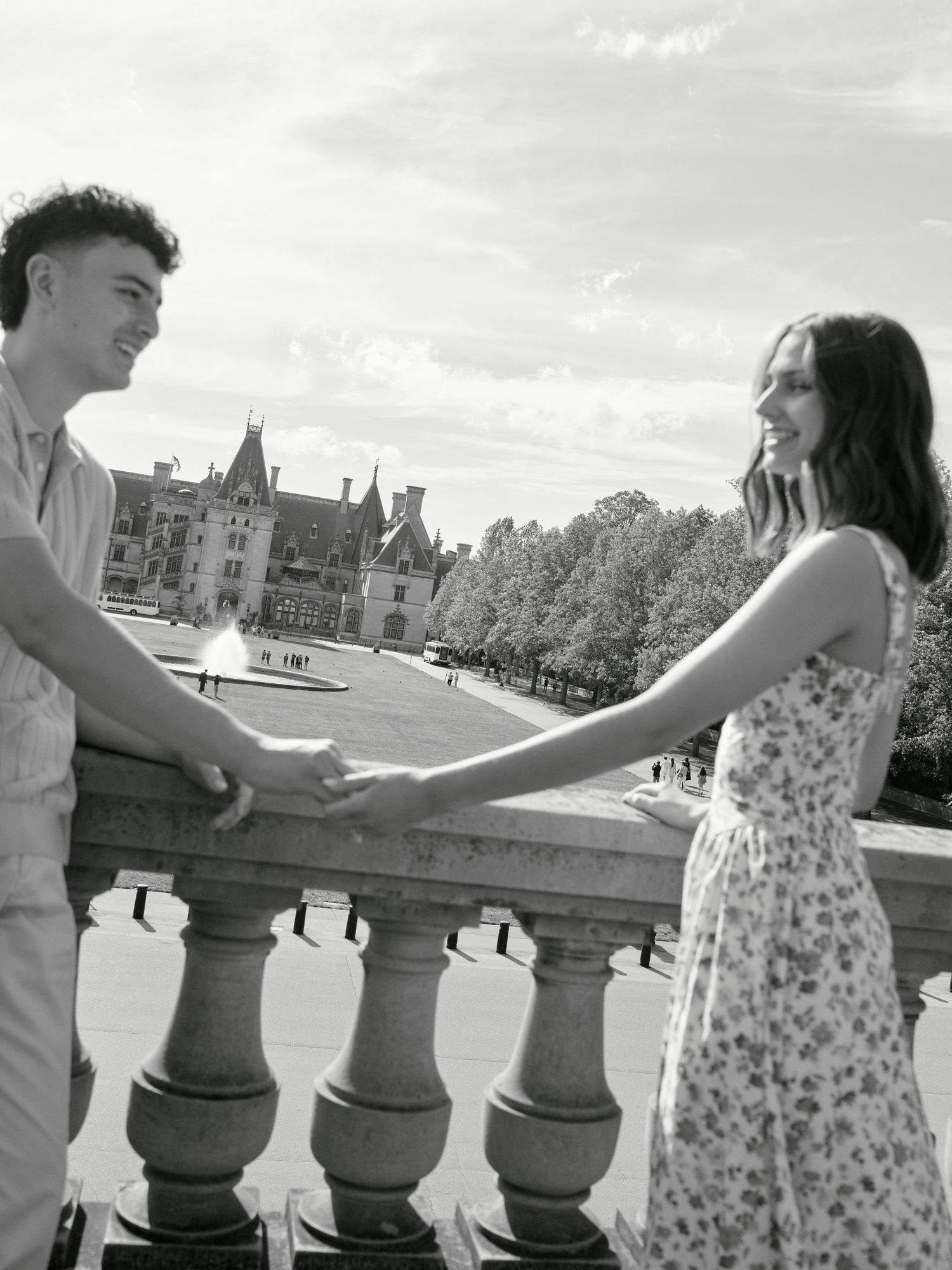 Black and white photo of a couple holding hands and smiling at each other near stone balustrade with a grand estate behind them.