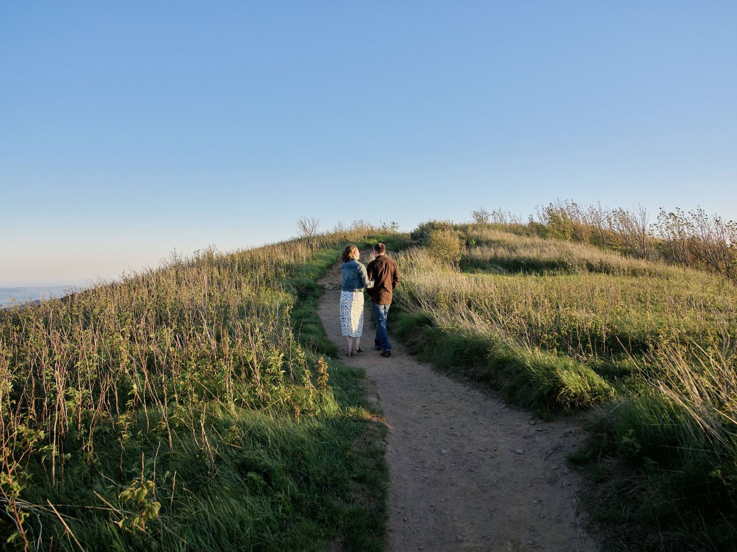 Couple walking hand in hand along a scenic coastal path surrounded by lush green grass under a clear blue sky.