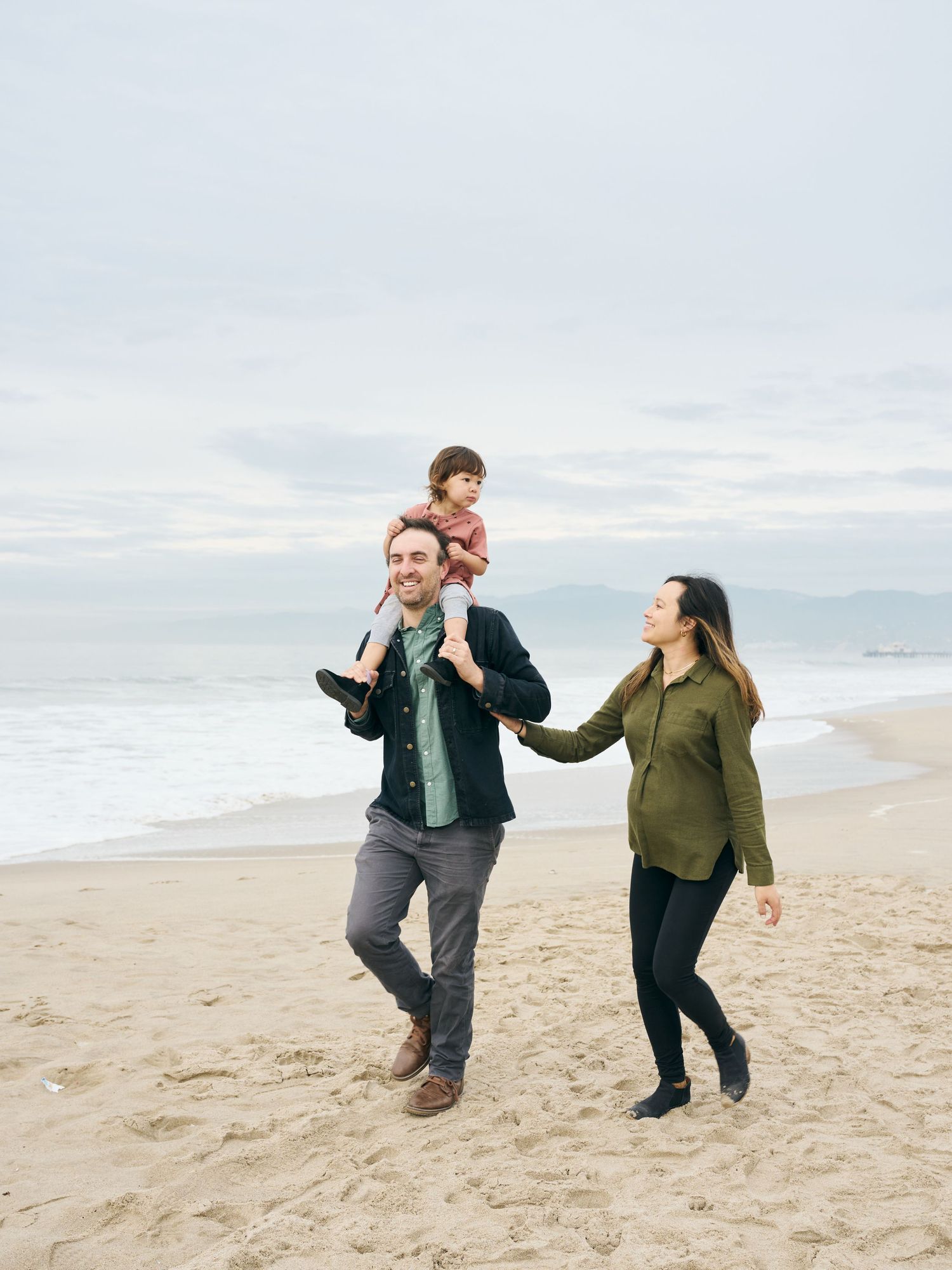 Family enjoying a walk together on a sandy beach with mountains in the distance on an overcast day.