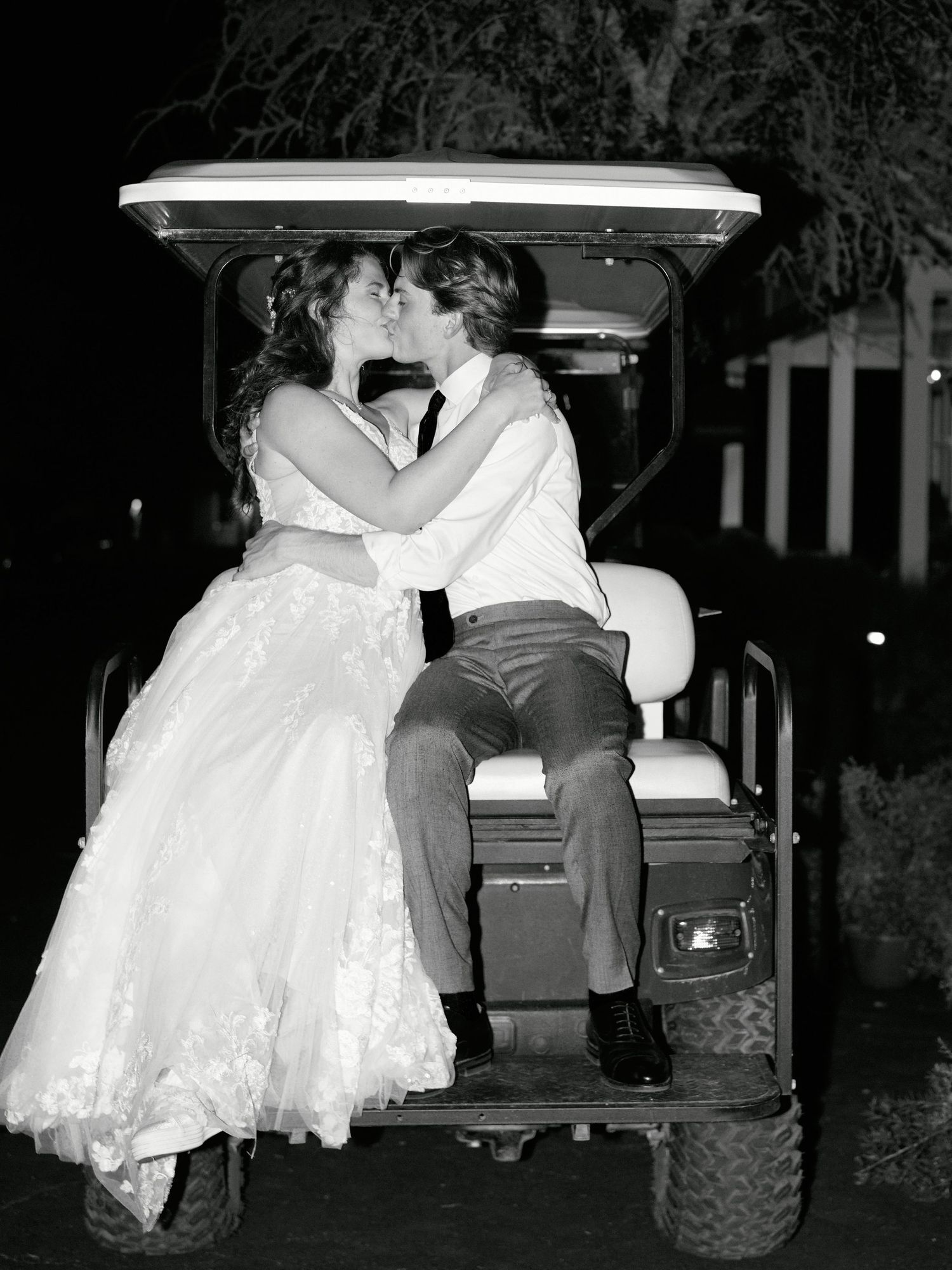 A romantic black and white photo of a couple in wedding attire sharing a kiss while seated in a golf cart at night.