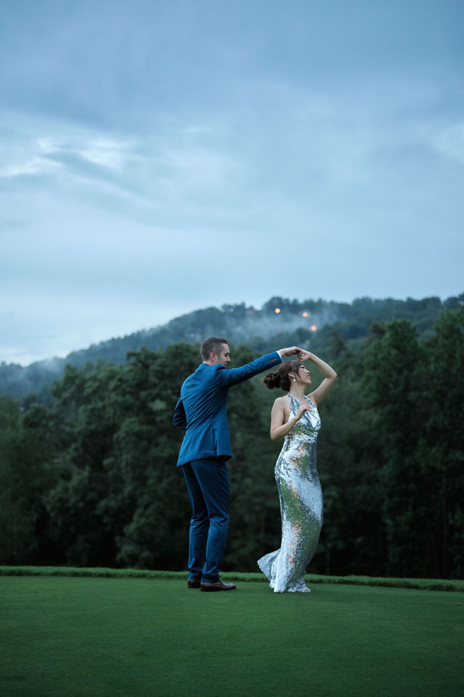 A couple dances playfully on a golf course with mountains and twilight sky in the background.