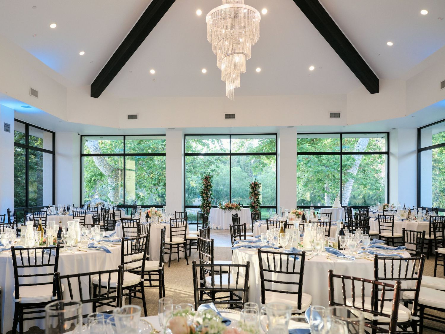 An elegant reception hall with exposed beams, crystal chandeliers and black chiavari chairs set for a wedding.