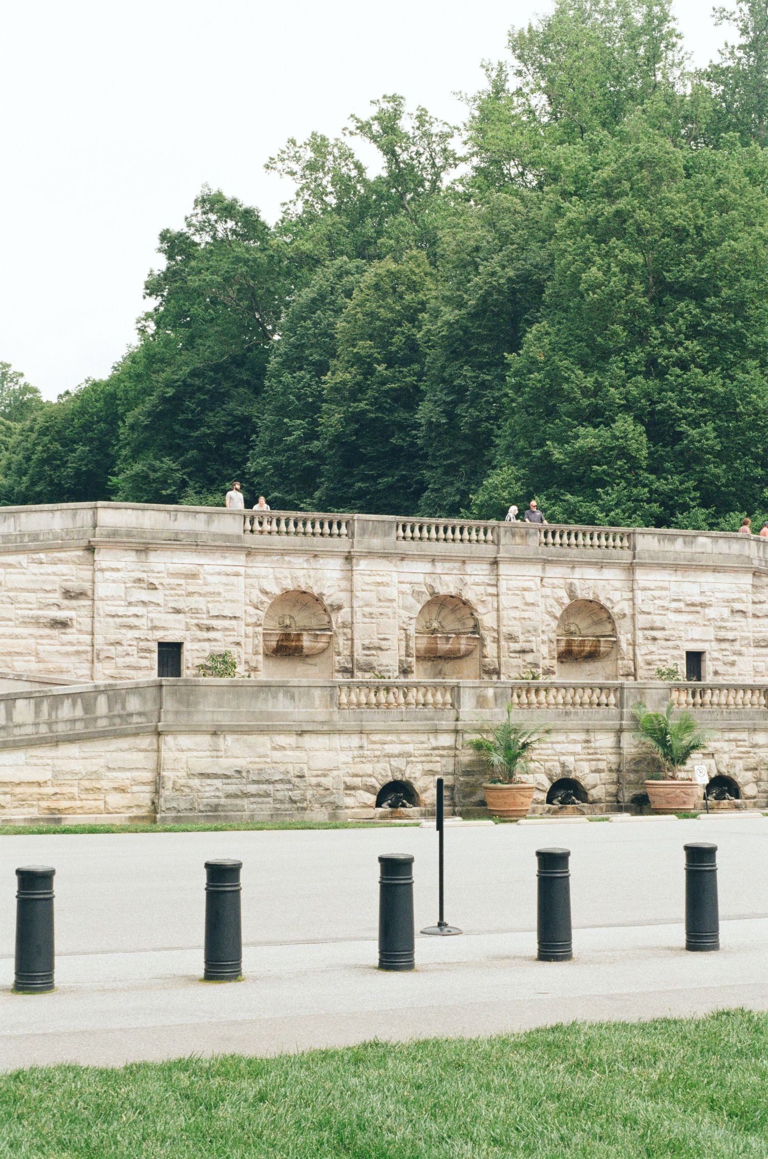 Stone wall with arched niches and decorative balustrade overlooks a grassy area in a park setting.