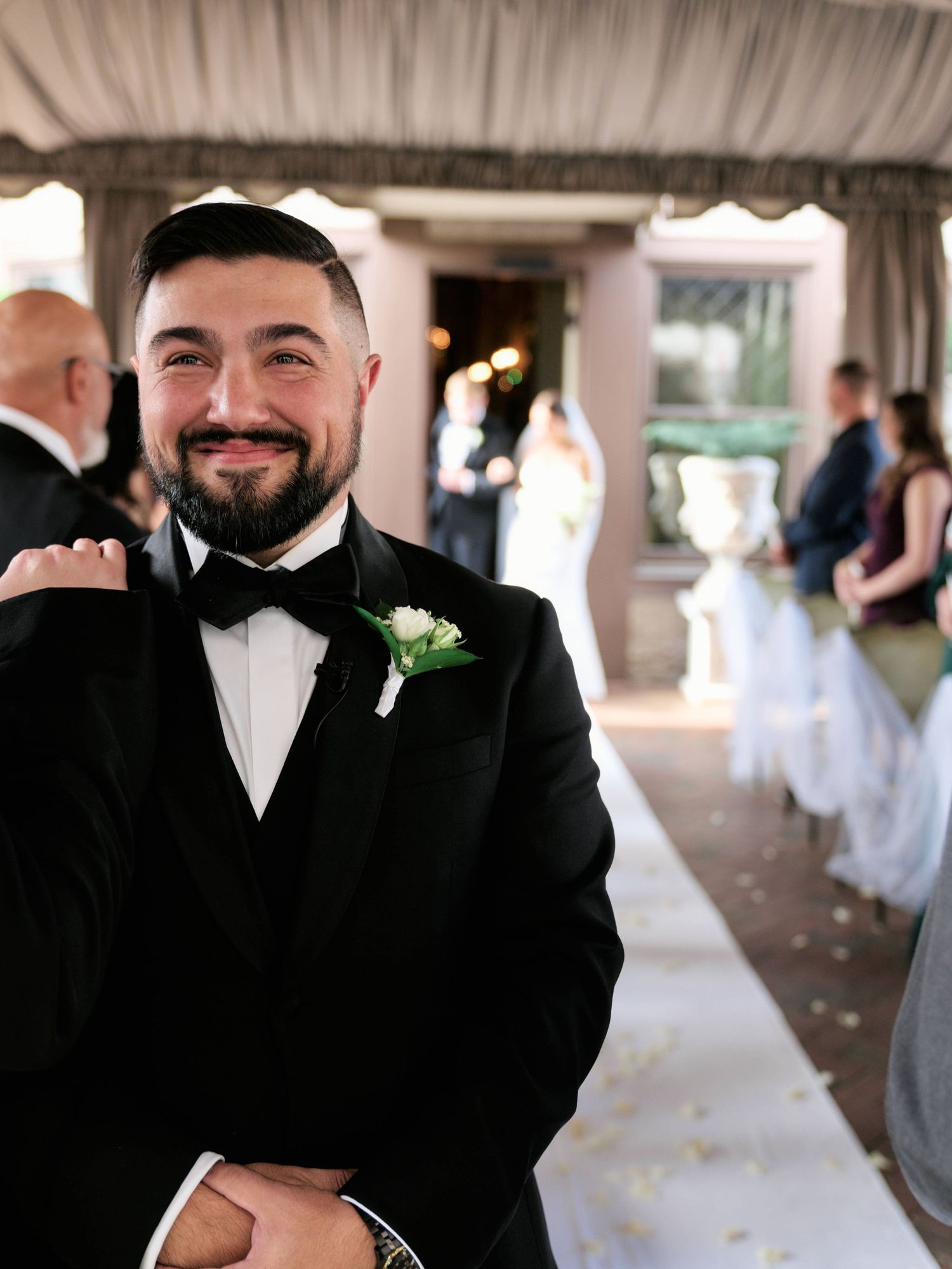 A groomsman in a black tuxedo and bowtie with a boutonniere smiles at a wedding venue with guests in the background.