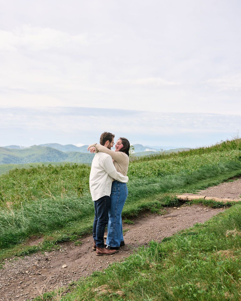 A couple embraces while standing on a mountain trail with rolling green hills and misty mountains in the distance.