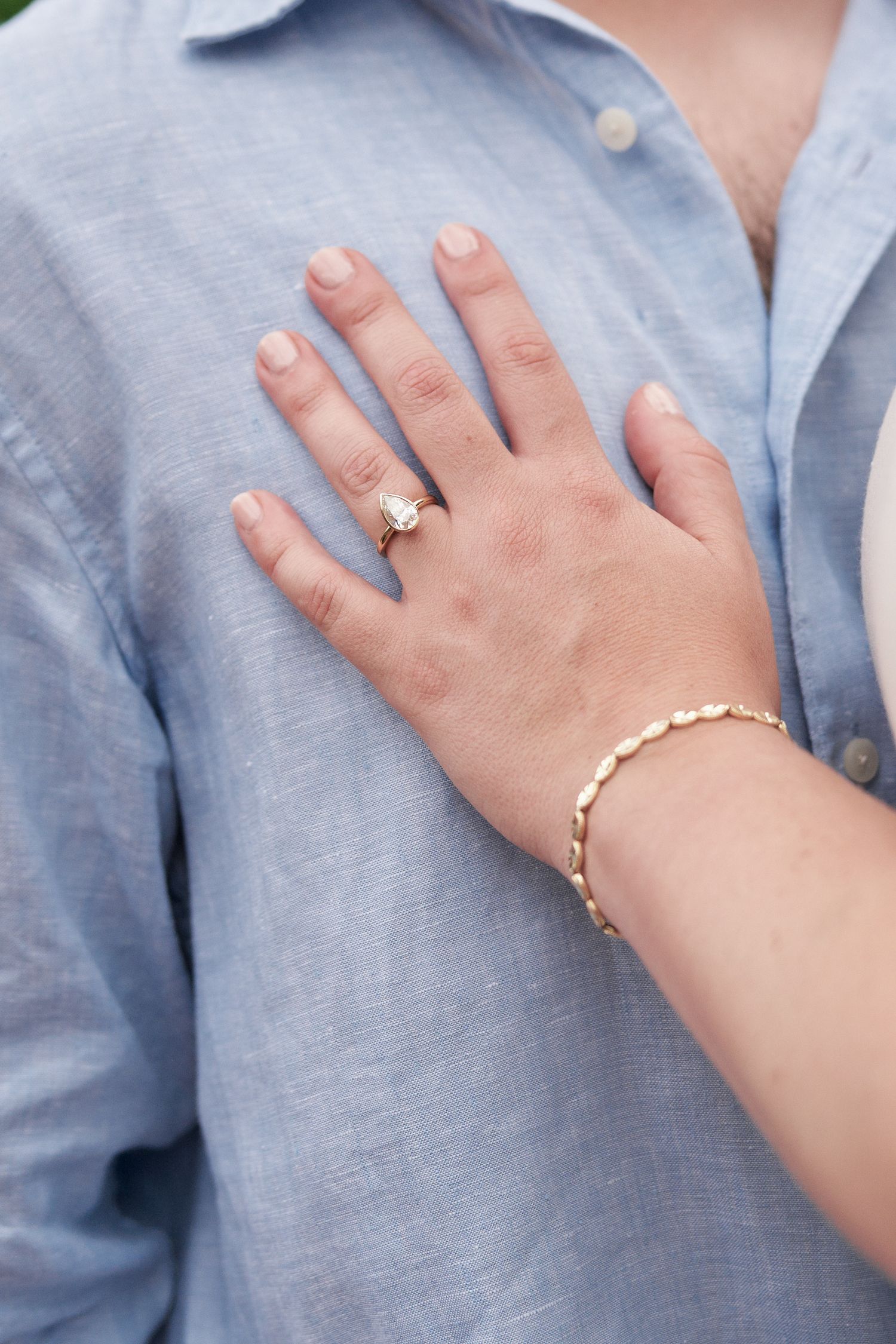 Close-up of an engagement ring and gold bracelet against a light blue shirt.