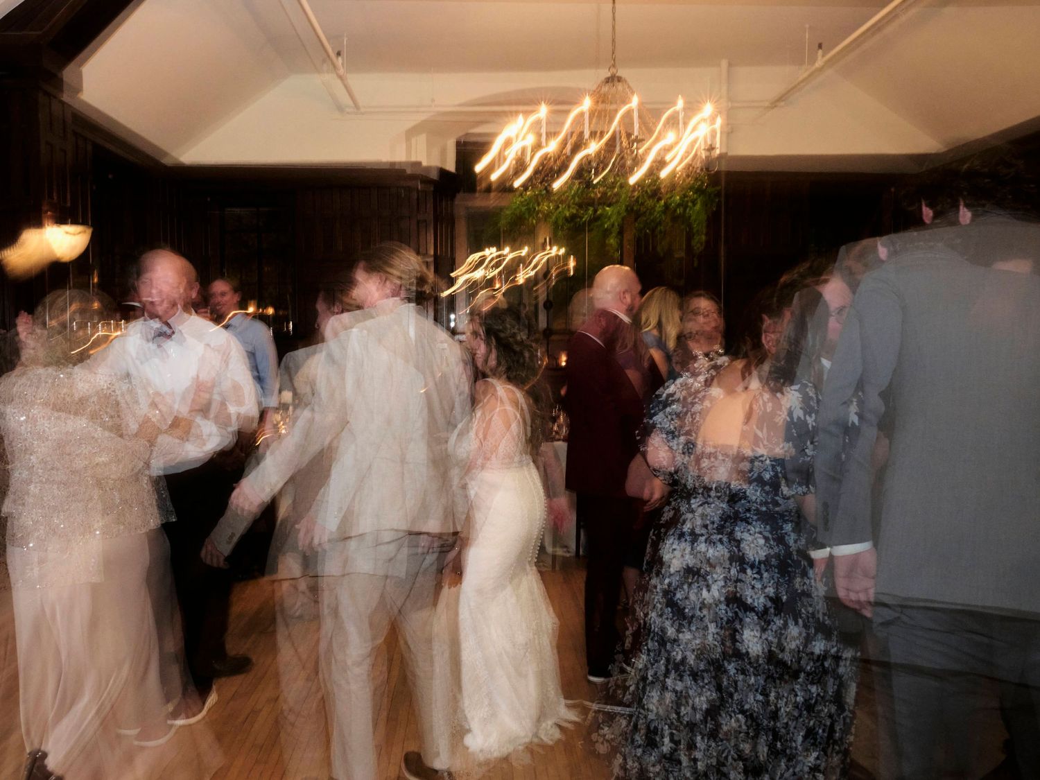 Blurred motion of wedding guests dancing at reception under chandelier and greenery.