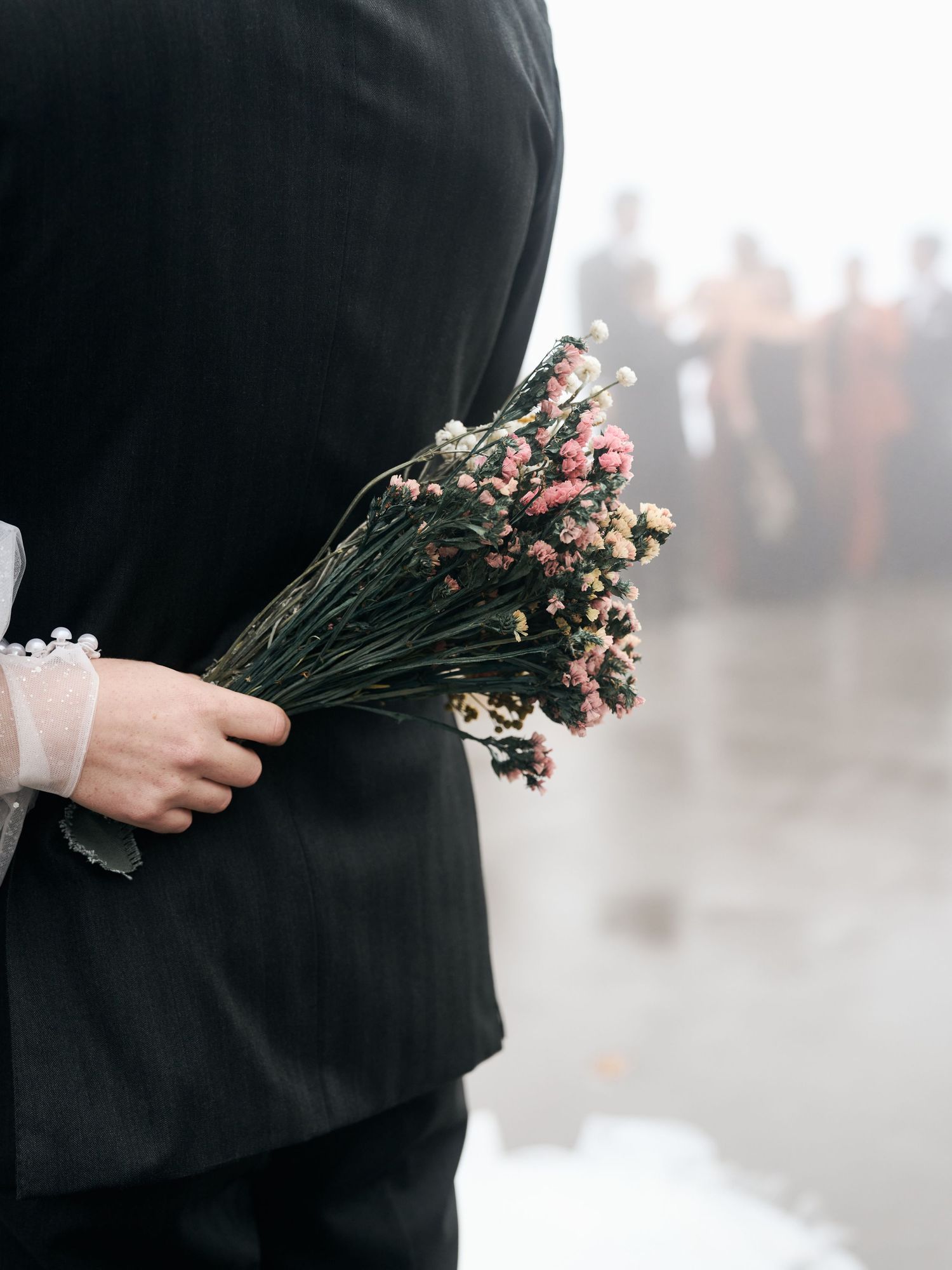 Person in formal attire holding delicate pink dried flower bouquet against misty background.