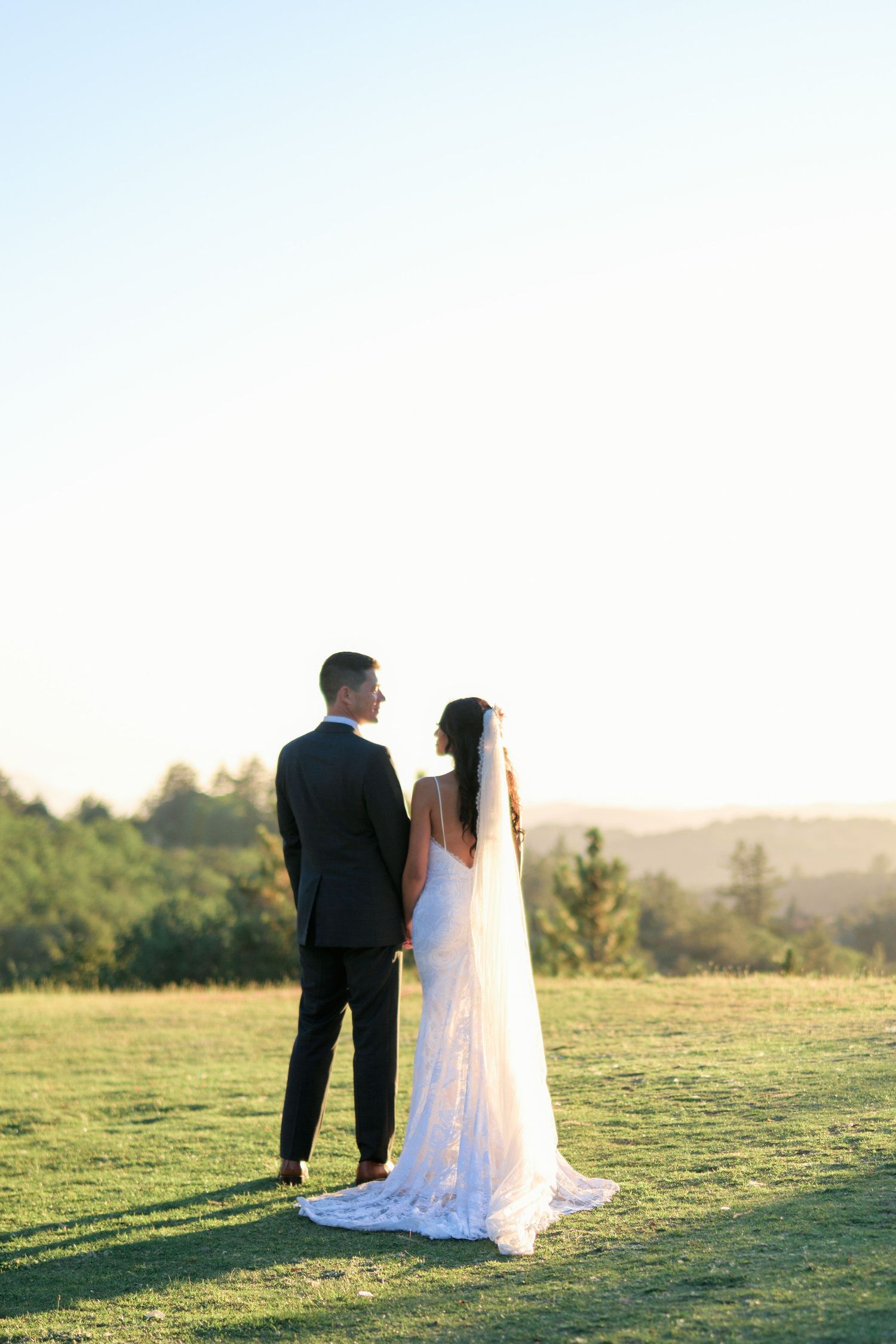 A wedding couple stands silhouetted against a bright sky on a grassy hilltop.