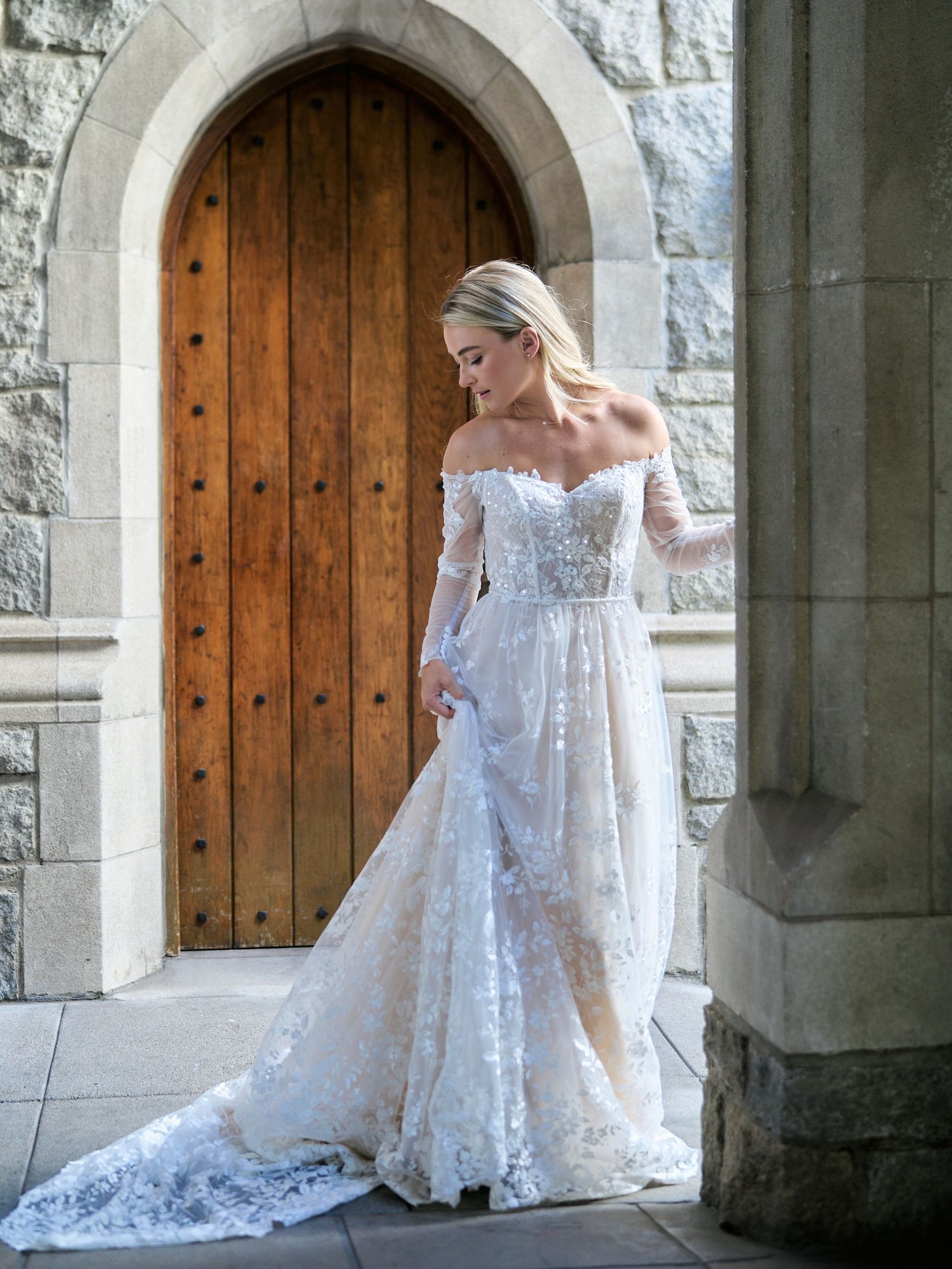 An elegant off-shoulder lace wedding dress with long sleeves photographed in a stone archway.