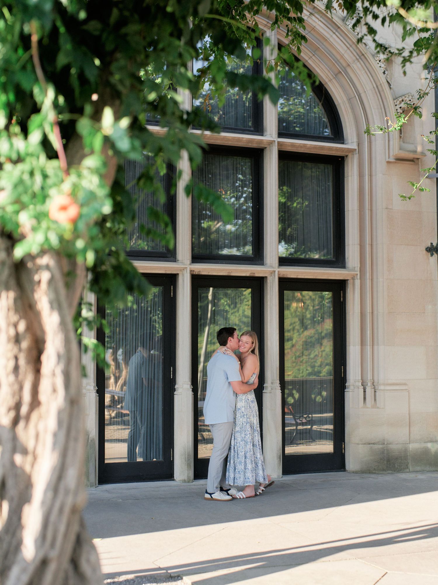A couple shares a romantic moment in front of arched windows along a stone building exterior with sunlit trees.