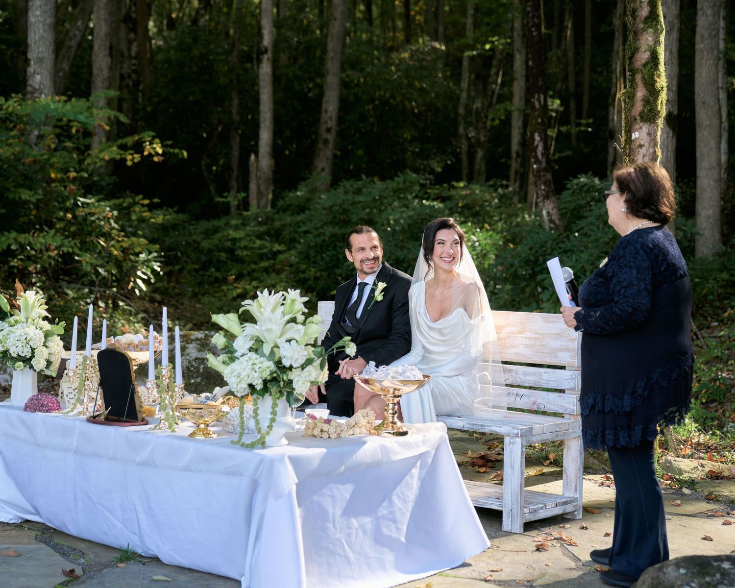 Outdoor wedding reception setup with long white tables and rustic wooden benches in a wooded forest setting.