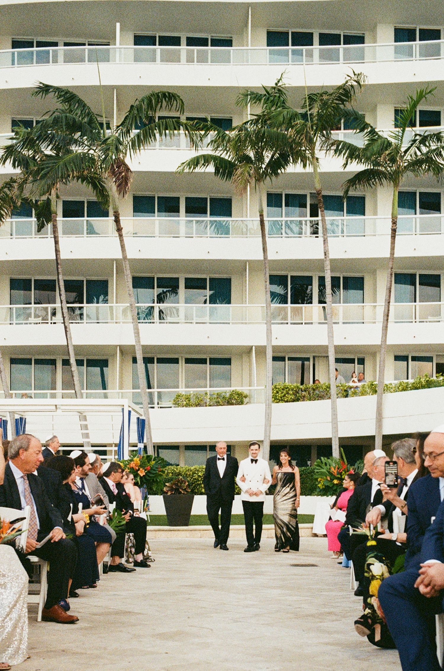 Outdoor wedding ceremony at a tropical hotel with palm trees, guests seated in aisle, couple at altar.
