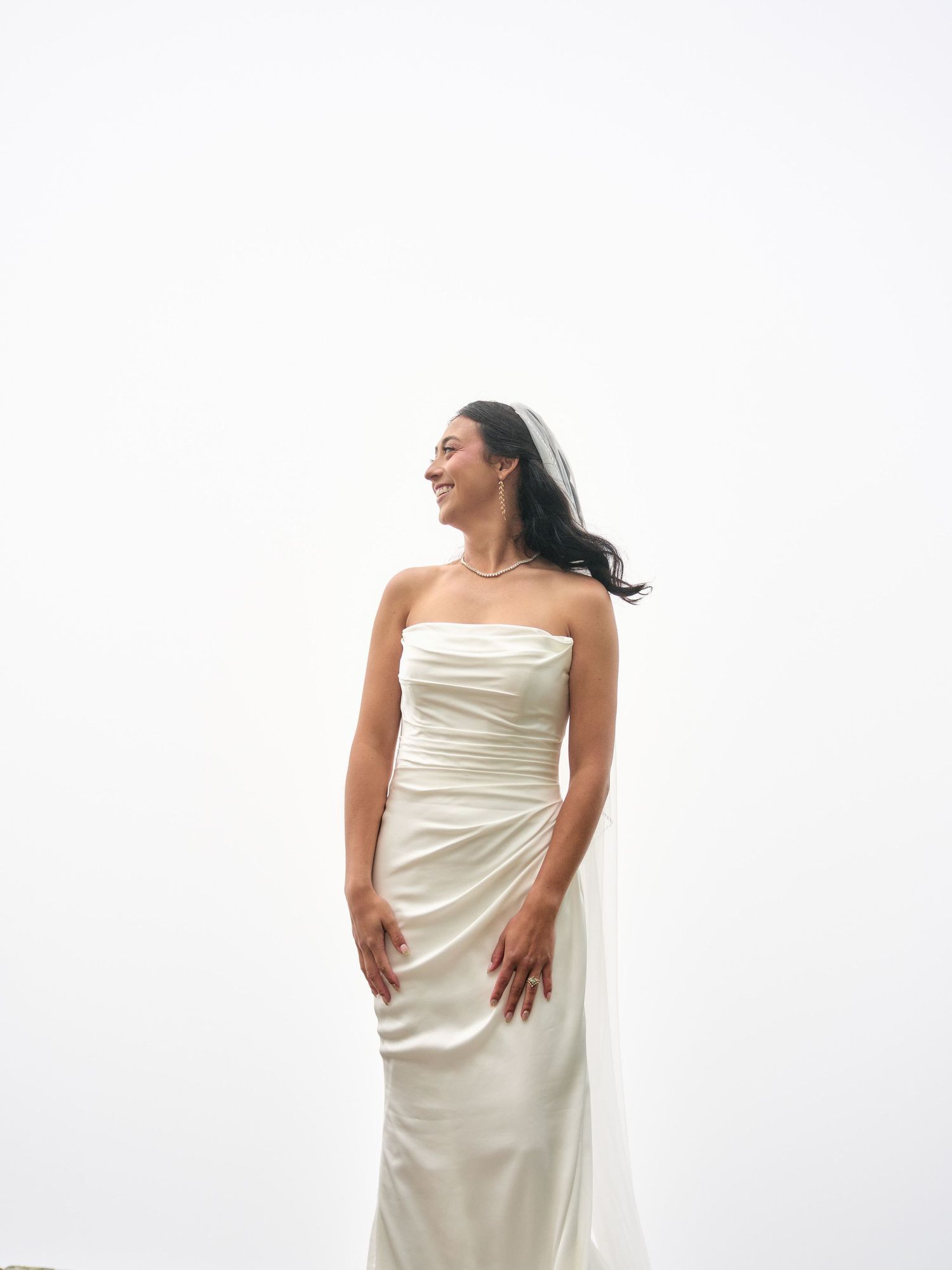 Smiling bride in a strapless ivory satin gown with ruching, wearing a veil, posing against a white background.