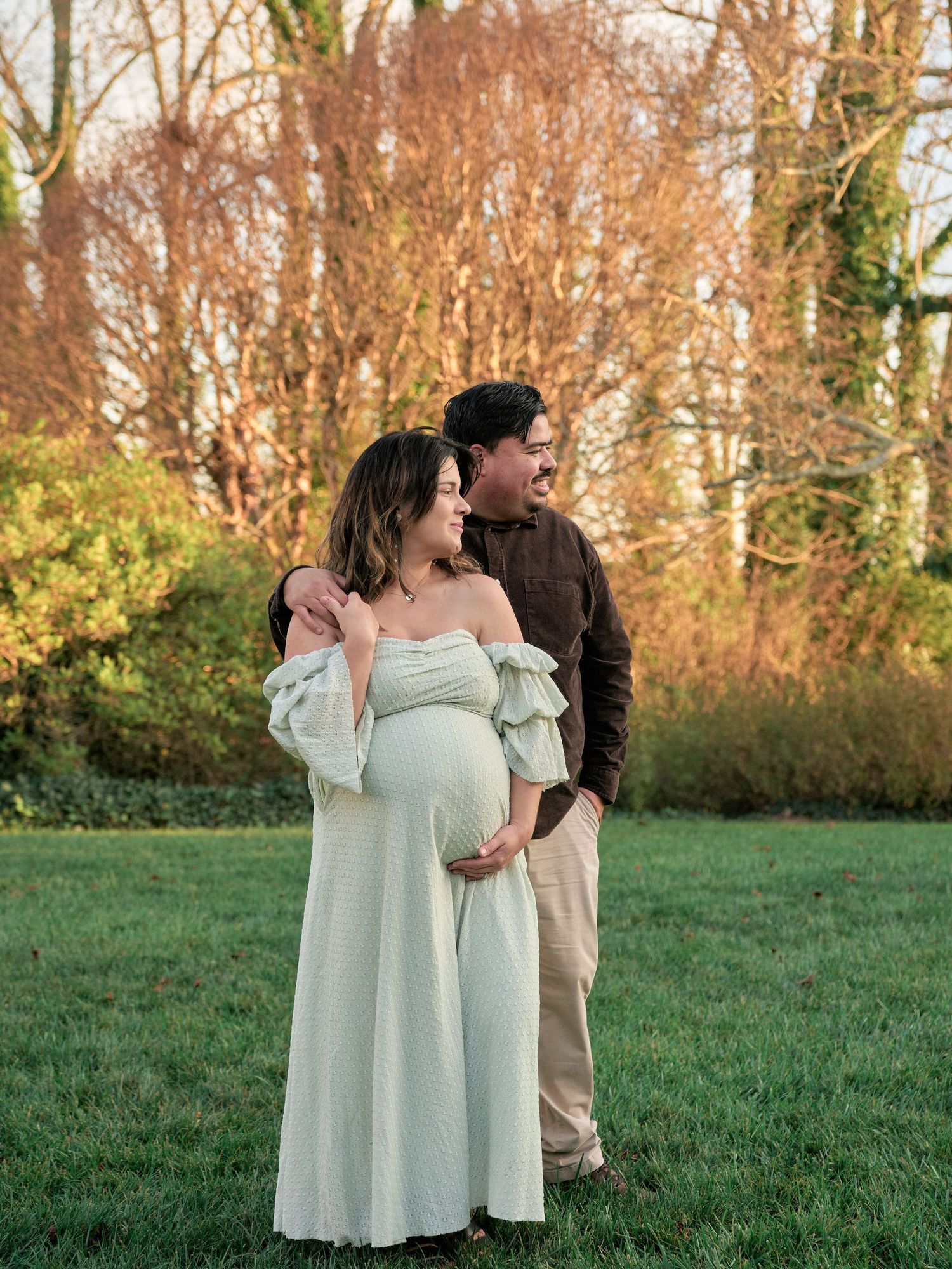 Pregnant couple posing outdoors in a lush green park, woman in mint off-shoulder maternity gown, man in brown top.