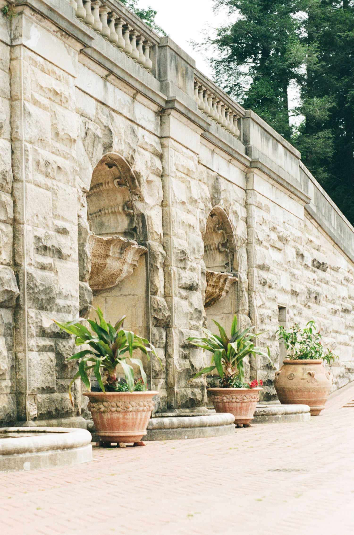 Stone wall with decorative architectural elements and potted plants along a curved pathway in a garden setting.
