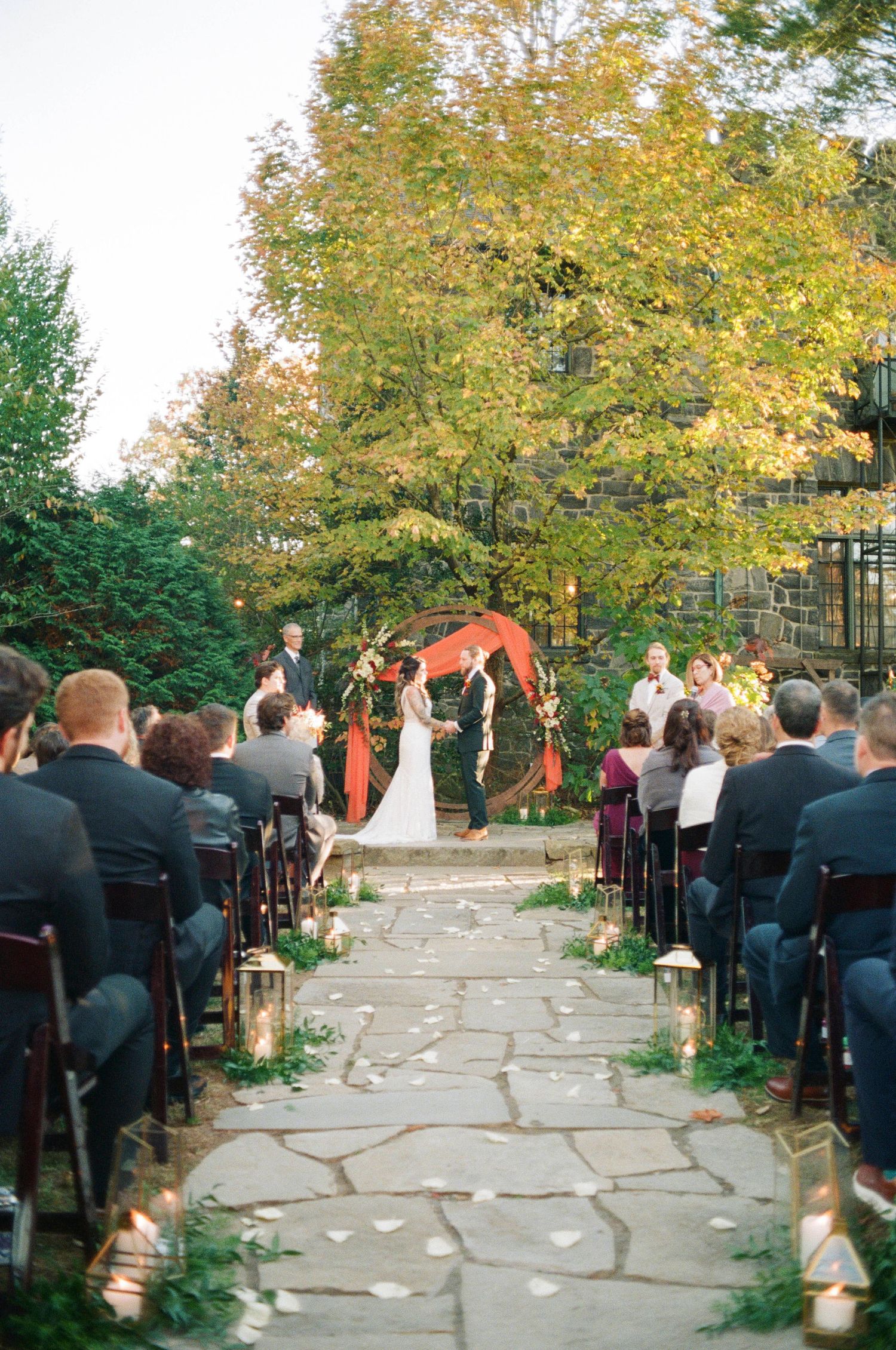 An outdoor wedding ceremony with guests seated along a stone path leading to an arch decorated in autumn colors.