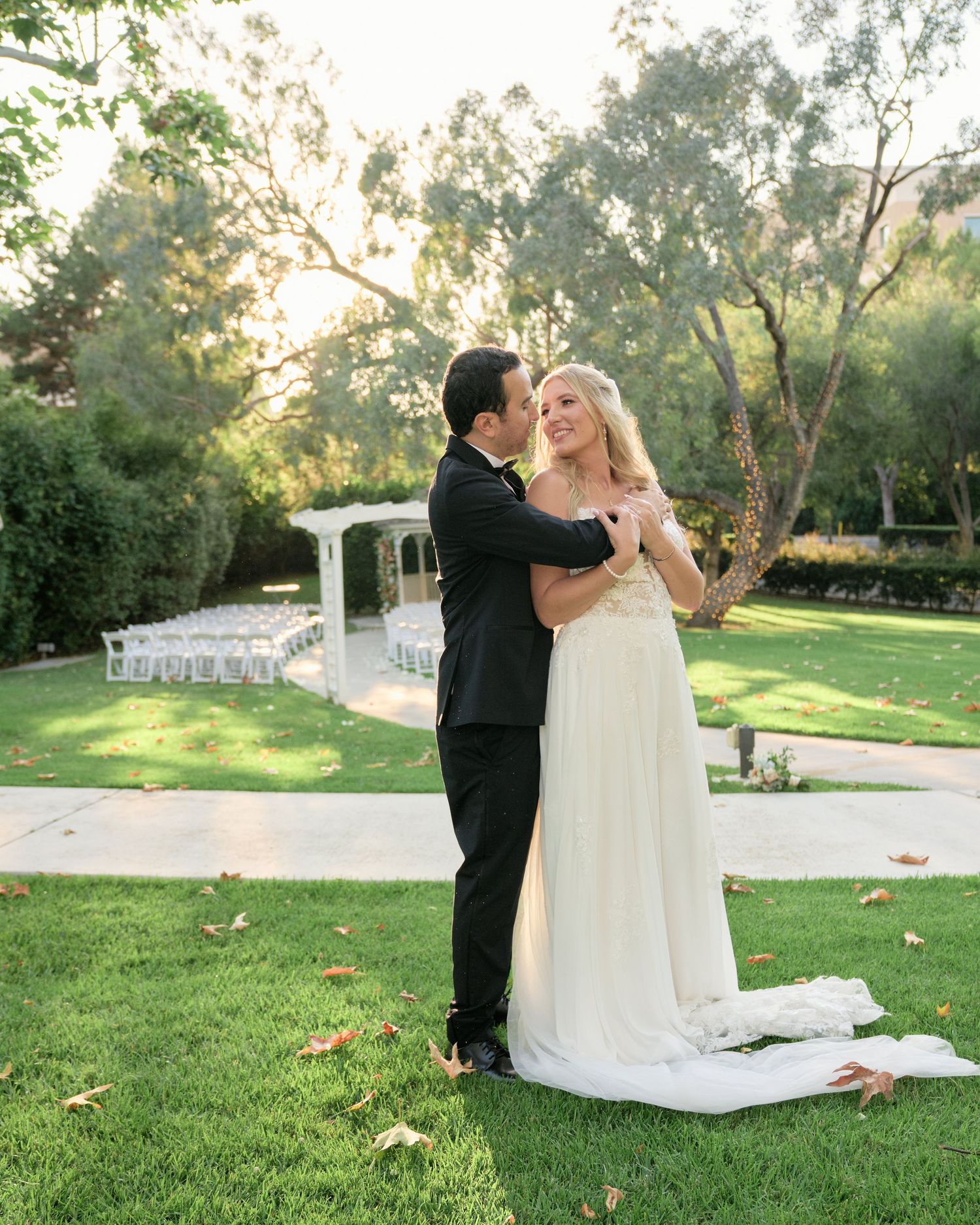 A romantic outdoor wedding scene with a couple embracing on a manicured lawn with a white gazebo in the background.