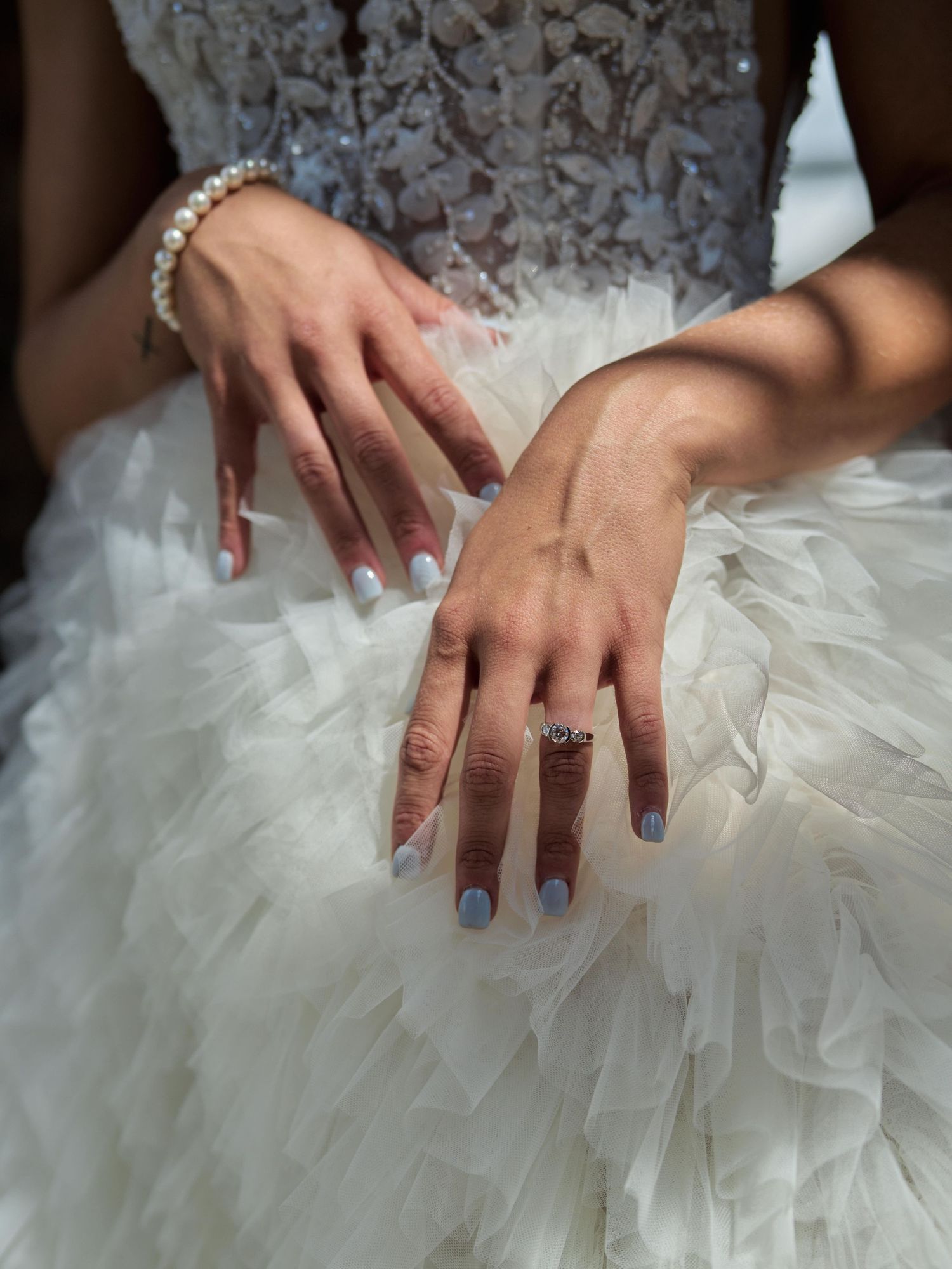 Close-up of bride's hands showing engagement ring, bracelet, and blue nail polish against tulle dress.