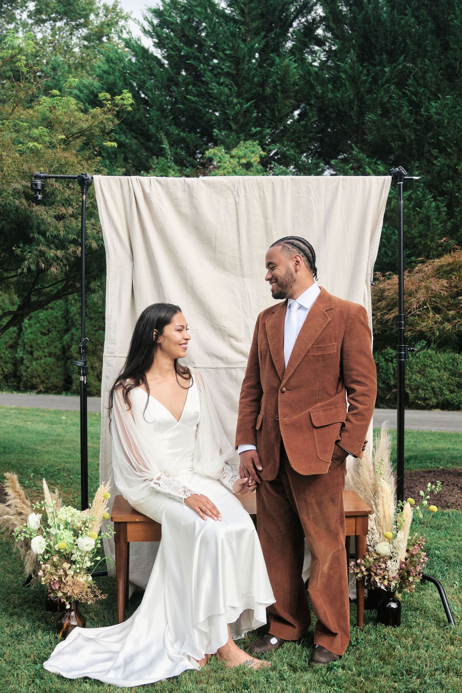 Joyful wedding couple poses casually against white backdrop in garden setting.