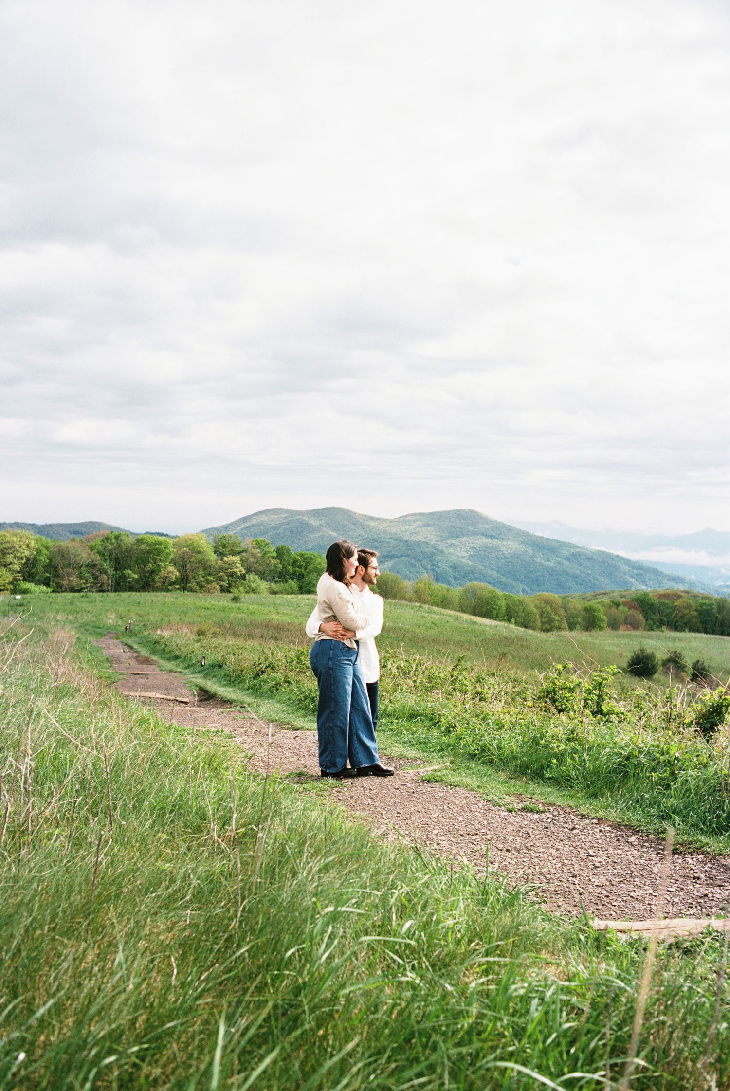 A couple stands embracing on a scenic mountain hiking trail surrounded by rolling green hills and meadows on a cloudy day.